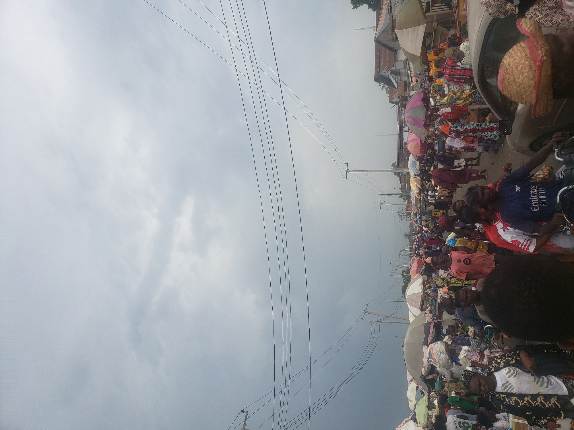This image captures a vibrant and densely populated outdoor market scene in Lokoja, Nigeria, under an overcast sky. The perspective is upward-looking, with a large portion of the frame dedicated to the cloudy sky, crisscrossed by numerous overhead power lines suspended from wooden utility poles.
The street level is bustling with a multitude of people, engaged in what appears to be a busy market. Individuals are seen standing, walking, and attending to various stalls. Many market vendors are using colorful umbrellas to provide shade over their goods, which are spread out on the ground or on makeshift tables. While specific items are difficult to discern, the impression is one of diverse wares for sale.
A silver car is parked on the right side of the street, and in the foreground, a person on a motorcycle is visible, wearing a blue t-shirt that reads "Emirates FLY BETTER." Another piece of text, "LAUNDRY BAG," is faintly visible on a white bag among the market items. Buildings with corrugated iron roofs line the background, contributing to the urban or semi-urban setting. The time of day appears to be daytime, under diffused light due to the heavy cloud cover. The scene exudes a sense of active commerce and community life.