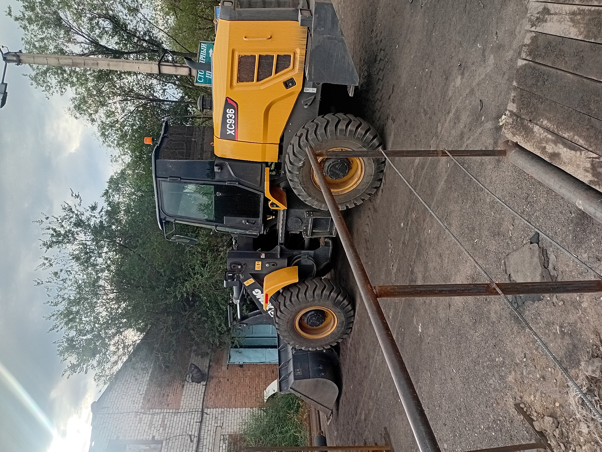 A yellow and black wheel loader, identified by the model number "XC936," is parked on a dusty, unpaved surface in an industrial or construction area. The loader has its bucket lowered to the ground. To the left of the loader is a distressed brick building with boarded-up windows and a blue metal door. To the right, a tall concrete utility pole with a street light is visible, along with a green sign that appears to have Cyrillic lettering. Overgrown green trees and vegetation are present in the background, and the sky is overcast. In the foreground, a metal railing with a cable running along its lower section suggests a barrier or walkway. The overall scene depicts an outdoor industrial setting, possibly within the city of Semey, Kazakhstan, given the context. There are no people visible in the image.