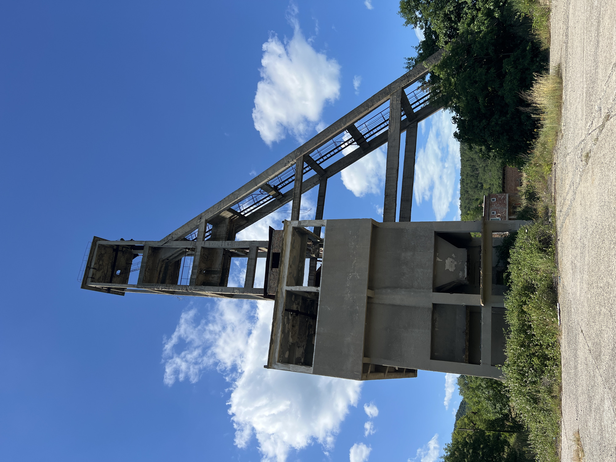 The image depicts a derelict industrial structure, likely a former processing plant or conveyor system, under a clear blue sky dotted with white clouds. The structure is made of concrete and appears to be a skeletal frame with open sections, suggesting it's abandoned or unfinished. A long, inclined conveyor belt structure dominates the scene, supported by a series of concrete pillars. Below and to the right, there are more concrete sections forming what might have been a processing or storage area.
The surrounding environment is lush with green trees and vegetation, indicating a rural or semi-rural setting. A dirt or gravel road runs along the right side of the frame, adjacent to the overgrown greenery. In the middle ground, partially obscured by trees, a small brick building with a few windows is visible, adding to the sense of a disused industrial site.
The lighting suggests it is daytime, likely mid-morning or afternoon, given the bright sunlight and clear sky. There is no activity or people visible in the image, emphasizing the sense of abandonment and stillness. The overall mood is one of decay and the passage of time, juxtaposed with the vibrant natural elements. No text is clearly legible in the image.