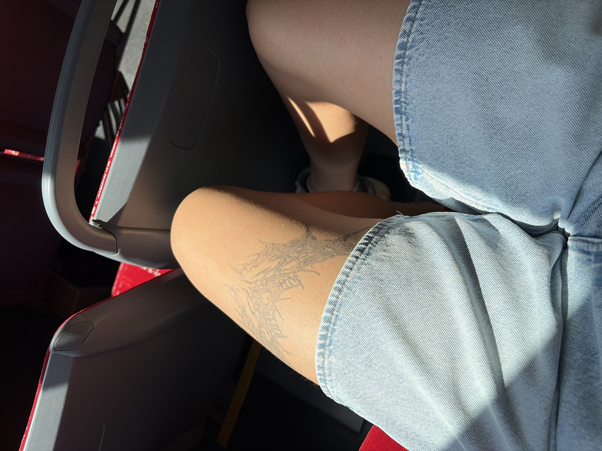 The image shows a close-up view of a person's thighs and denim shorts, taken from a seated position. The person has a tattoo on their left thigh, which appears to be a spiderweb design. Sunlight streams in from the right, casting shadows across the scene. The background reveals the interior of what appears to be a bus or train, with red seats and grey plastic elements. The specific location in the city of Nitra, Slovakia cannot be determined from the visual information provided.