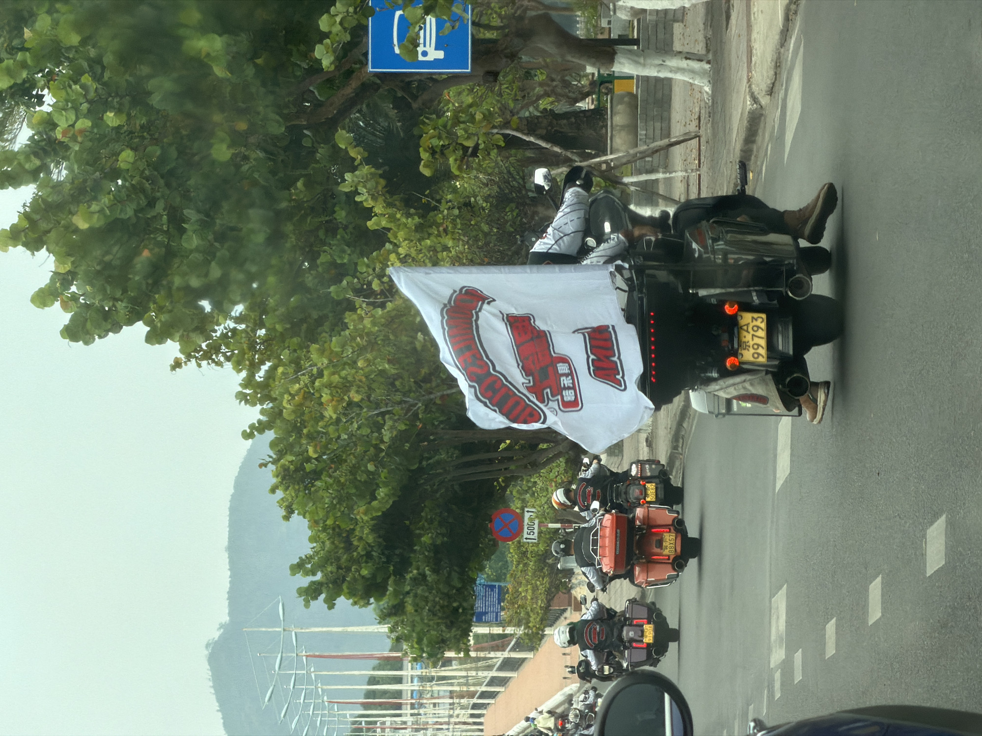 The image shows a motorcade of motorcycles traveling along a road in the Nha Trang Ward, Vietnam. The lead motorcycle has a white flag with a red logo. The motorcycles are traveling in a straight line, and there is a person riding each motorcycle. Trees line the side of the road, and there is a sign marking parking for electric vehicles. In the background, masts of sail boats and mountains are visible.