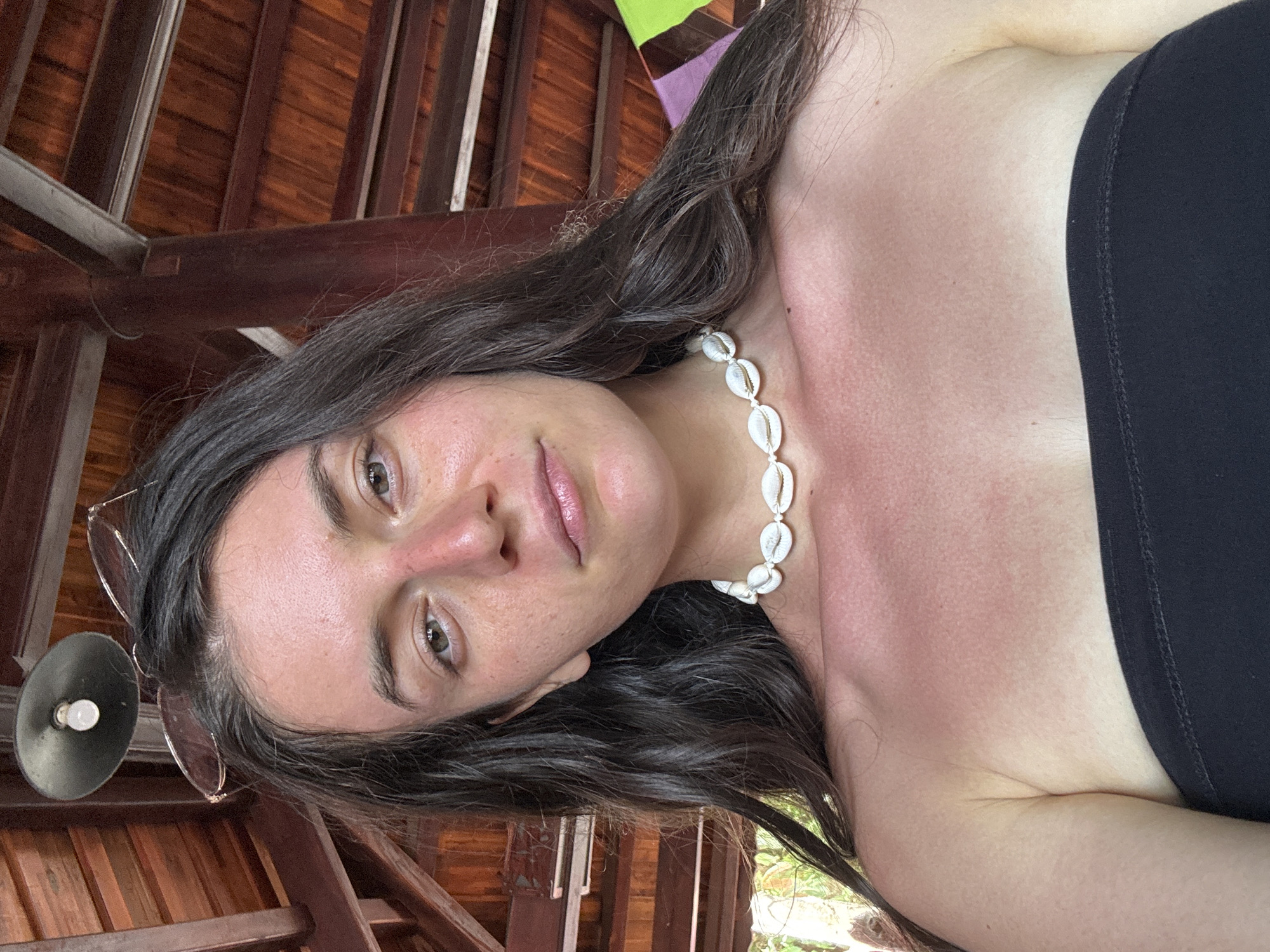 The image shows a young woman indoors in Mũi Né, Vietnam. She is wearing a black strapless top and a white seashell necklace. Her skin appears sunburnt. She has long, dark hair and is looking directly at the camera. A ceiling and wooden beams are visible above her, and a lamp is hanging nearby.