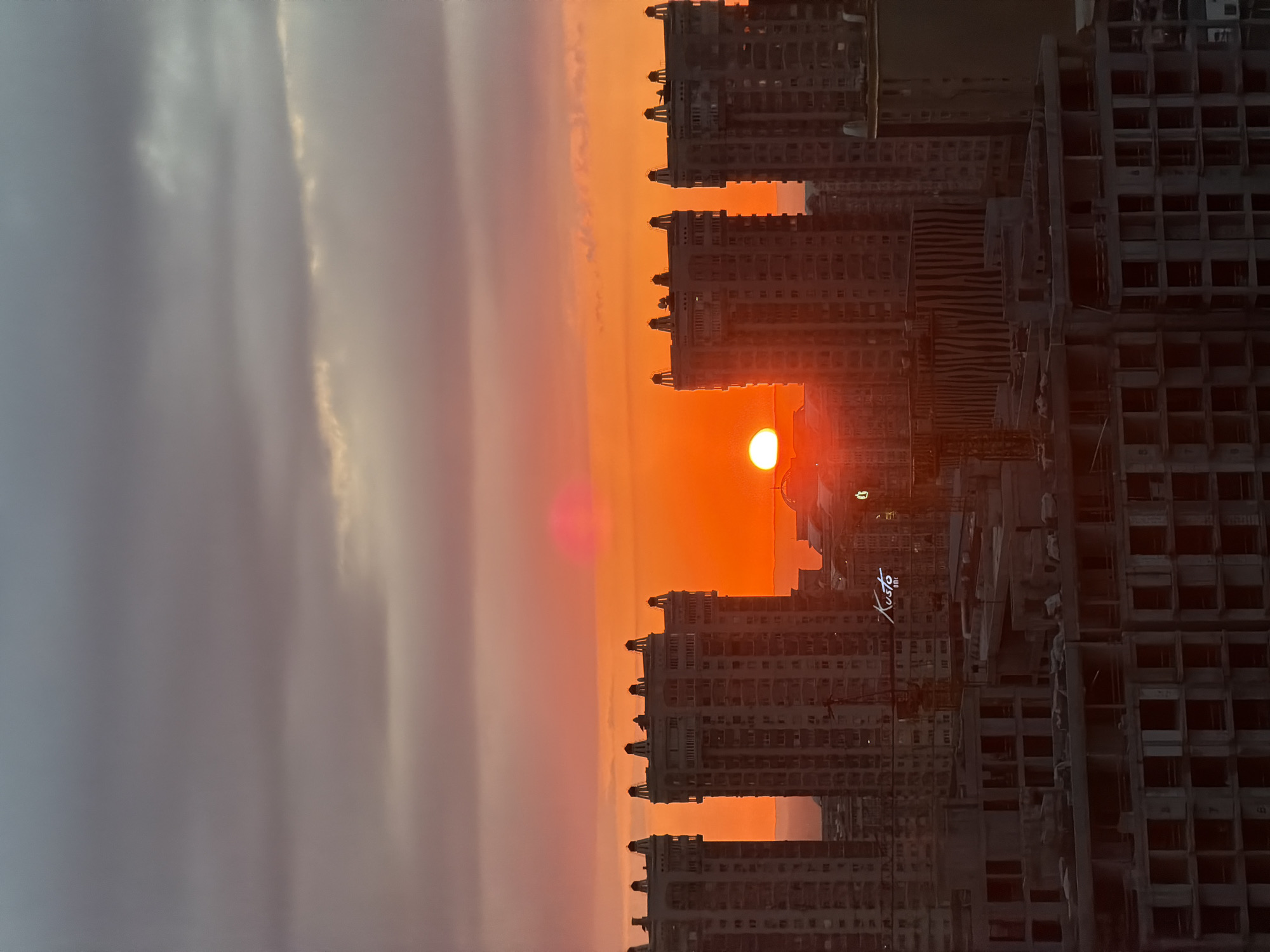 The image depicts a sunset over a cityscape in Almaty, Kazakhstan. The sun is positioned low on the horizon, appearing as a bright orange orb amidst the orange-hued sky. The sky transitions from gray clouds on the left to shades of orange and pink near the sun. Silhouetted against the setting sun are several high-rise buildings, some of which are still under construction. A sign with the name "Kusto" is visible on one of the buildings.