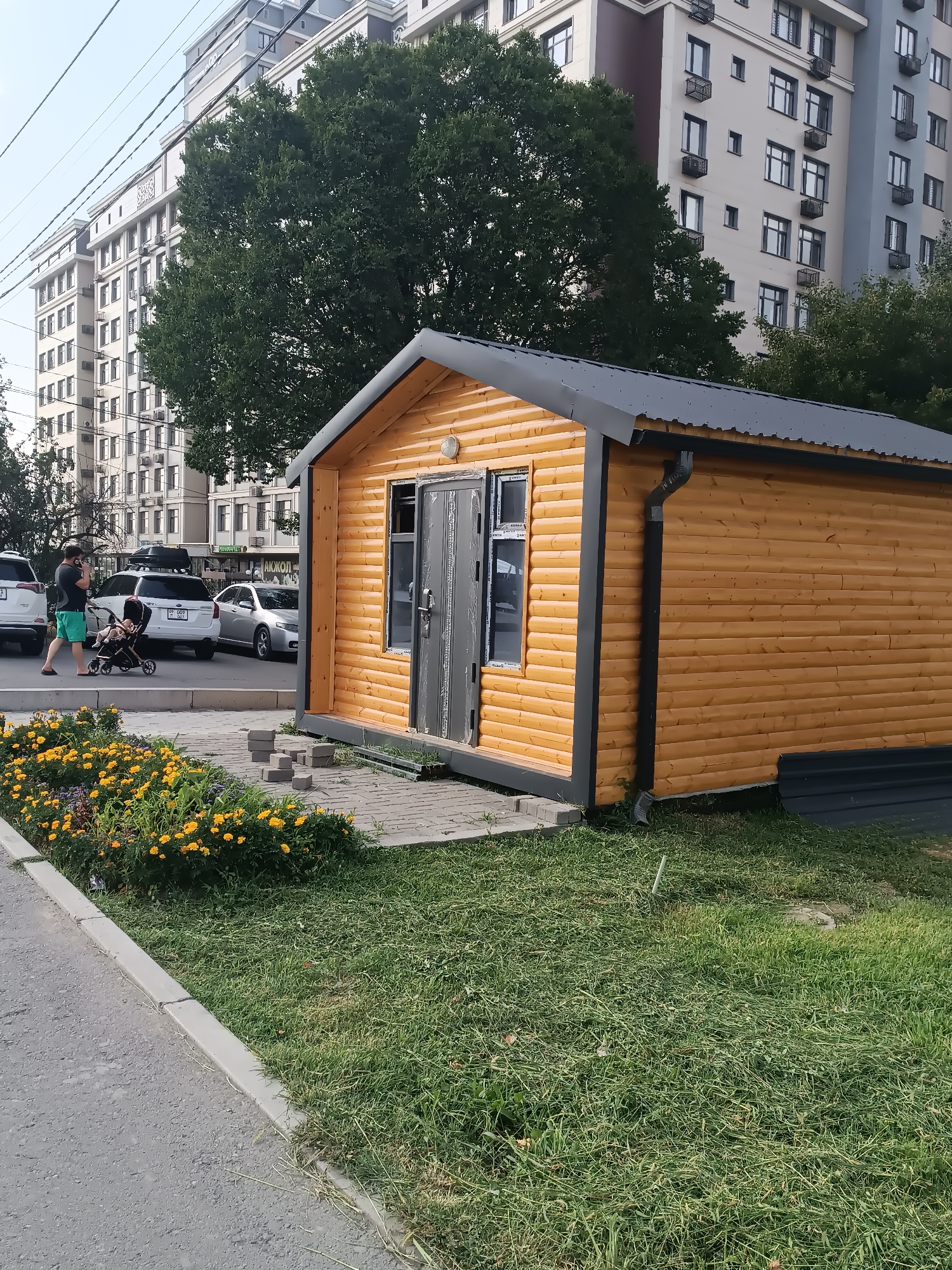 An outdoor, eye-level shot captures a small, newly constructed wooden shed with a dark grey, corrugated metal roof. The shed, situated on a paved area, has a light brown wooden facade with dark grey trim around the windows and doors. A single window with grey framing is visible on the side, and a double door with similar framing is on the front. A dark grey downspout is attached to the side of the shed, directing water to the ground.
In front of the shed, a patch of vibrant yellow flowers and green foliage lines a paved walkway. A man wearing green shorts and a dark t-shirt is pushing a stroller with a child in it, walking on the paved area to the left of the shed. Behind them, several parked cars are visible, including a white SUV and a silver sedan.
The background features multi-story apartment buildings, some with a light beige facade and others with a bluish-grey exterior. Tall green trees are interspersed between the buildings. Overhead, a network of power lines crisscrosses the sky. The overall scene appears to be a residential or mixed-use area in Bishkek City, Kyrgyzstan, possibly a new development or a public space undergoing renovation. The presence of the unfinished shed suggests ongoing construction or a recently installed feature.