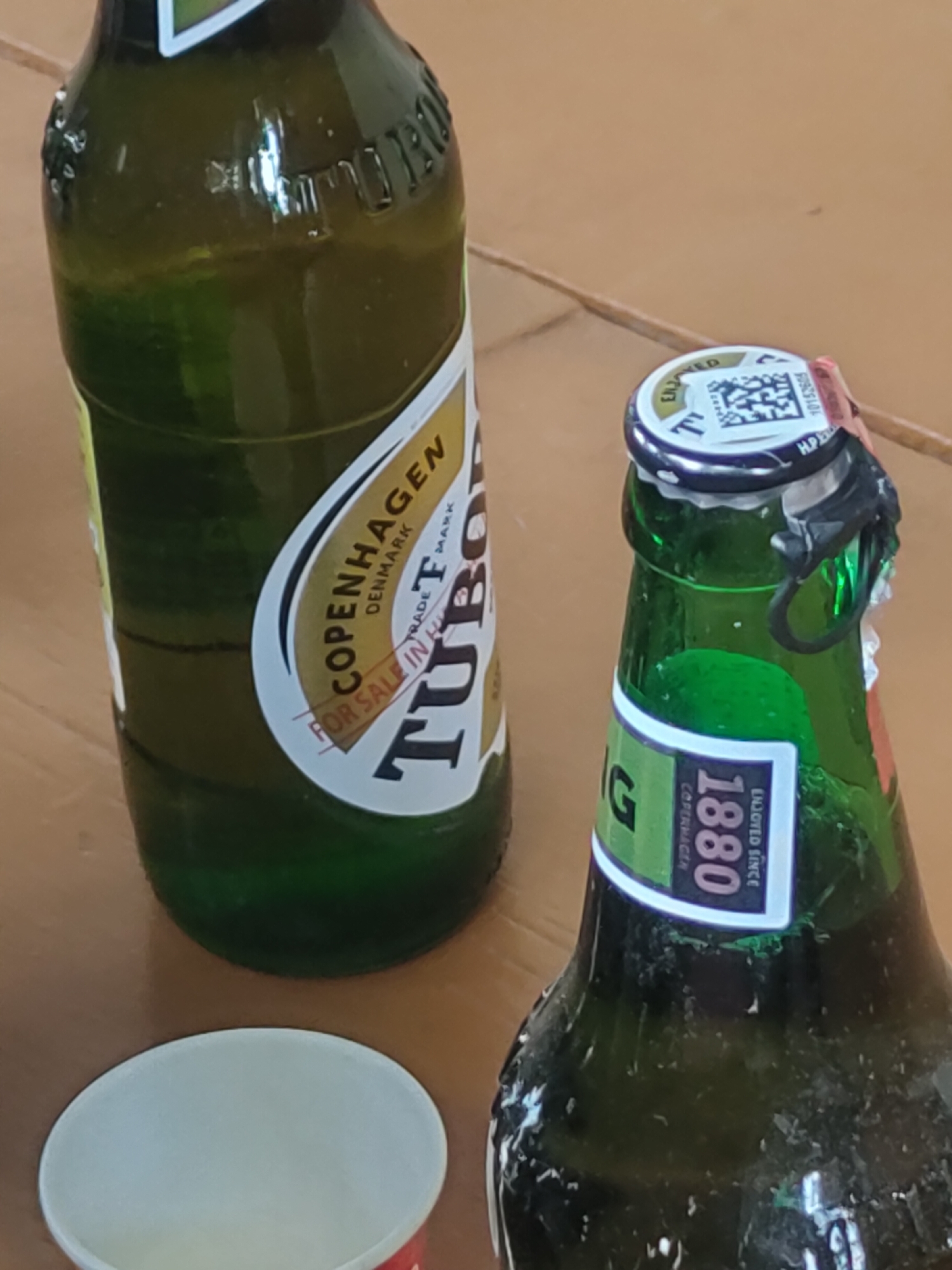 The image shows two green glass bottles of Tuborg beer placed on a light brown wooden surface. One bottle is in the foreground on the right and is upright. Its cap has a QR code printed on it and a red and silver seal partially covering the cap. A label on this bottle reads "G COPENHAGEN 1880 ENJOYED SINCE". The other bottle is on the left side of the image, also upright, and slightly out of focus. It has a white label with yellow text that reads "COPENHAGEN DENMARK TRADE MARK FOR SALE IN HK TUBO". The bottles appear to be full or mostly full. A white paper cup with a red rim is visible in the bottom left corner of the image. The scene is indoors, on a wooden table. The lighting suggests it is daytime. There are no people visible in the image. The image does not provide any information about the city of Burwa, India.