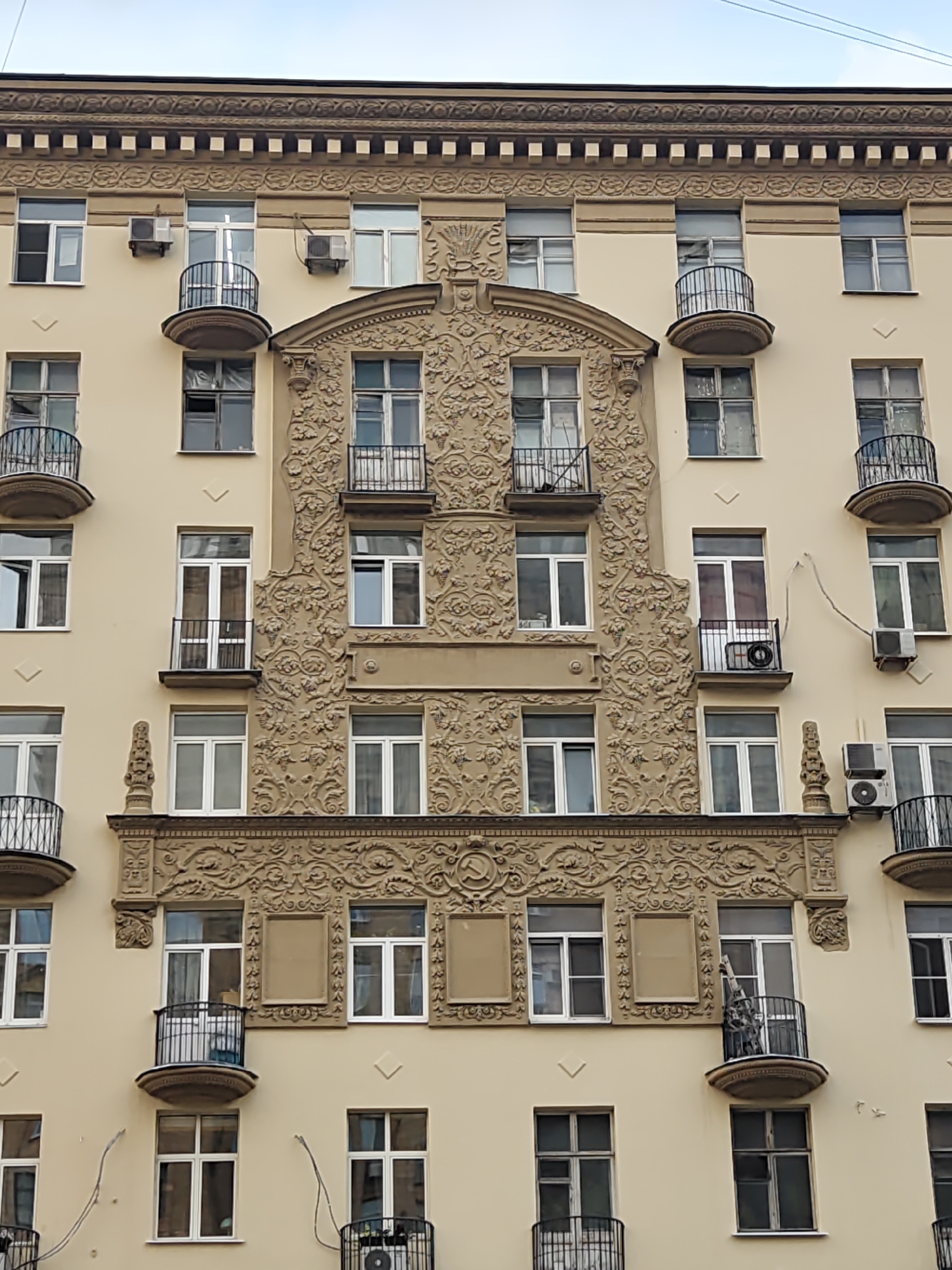 This is a low-angle, full-frame, eye-level exterior shot of a pale yellow apartment building in Moscow, Russia. The building features an ornate, light brown, stucco facade with repeating decorative elements. Rows of windows with white frames are evenly spaced across the building's surface, interrupted by small, curved balconies with black metal railings. Several windows have air conditioning units mounted beneath them, and some of the balconies also have AC units. The building exhibits signs of wear, with some windows appearing dirty or damaged. A prominent decorative band runs horizontally across the upper portion of the building, featuring intricate carvings of floral motifs and geometric patterns. Two Soviet-era symbols, a hammer and sickle, are visible within this decorative band, one on the left and one on the right. The sky is overcast and appears pale gray.