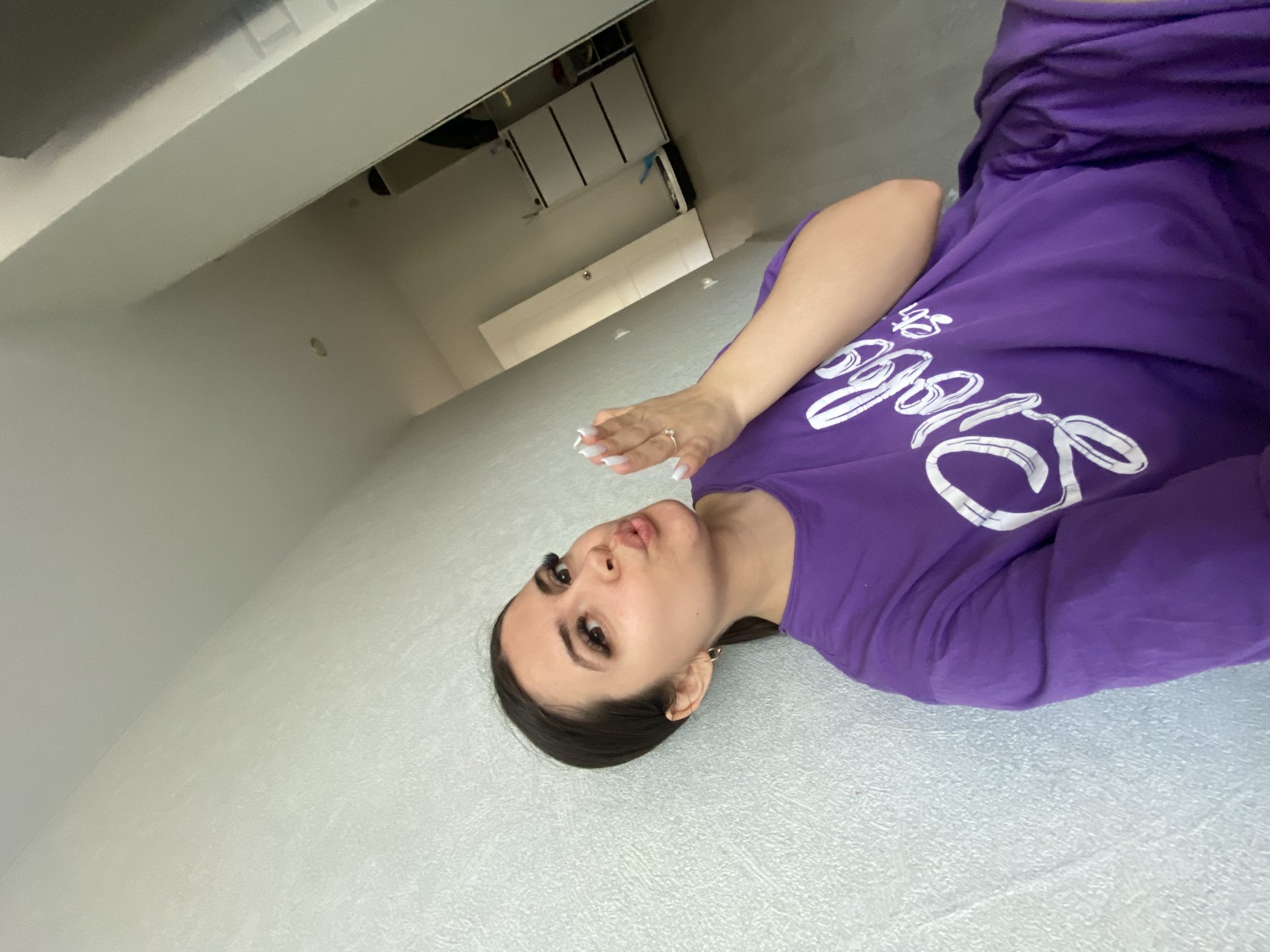 A woman is lying on a gray textured floor wearing a purple long-sleeved shirt with white lettering. She has long dark hair pulled back, dark eyebrows, and eyelash extensions. Her left hand is raised, showing long white nails and a ring. Behind her is a gray wall. In the background, there is a white door and white cabinets. This image was taken inside a building in Chaikovsky, Russia.