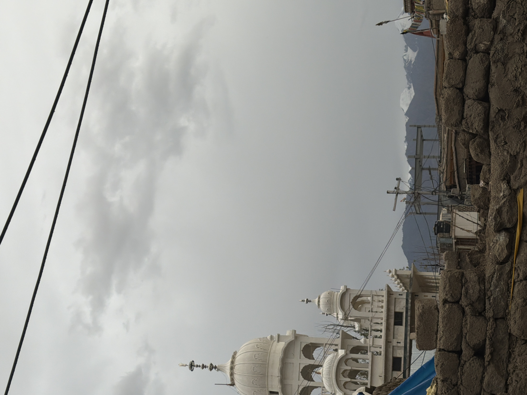The image depicts a view of a Gurdwara, a Sikh place of worship, in the city of Leh, India, under an overcast sky. The Gurdwara is a white, multi-tiered structure with domes and arched windows, characteristic of Sikh architectural style. In the foreground, a low wall constructed of rough, mud-brick materials blocks the lower part of the view, suggesting ongoing construction or a humble neighborhood. Wires are strung across the sky, and in the background, snow-capped mountains are visible, indicating the mountainous terrain surrounding Leh. The overall atmosphere is somber due to the cloudy weather. There are no people visible in the frame, and the scene is static.