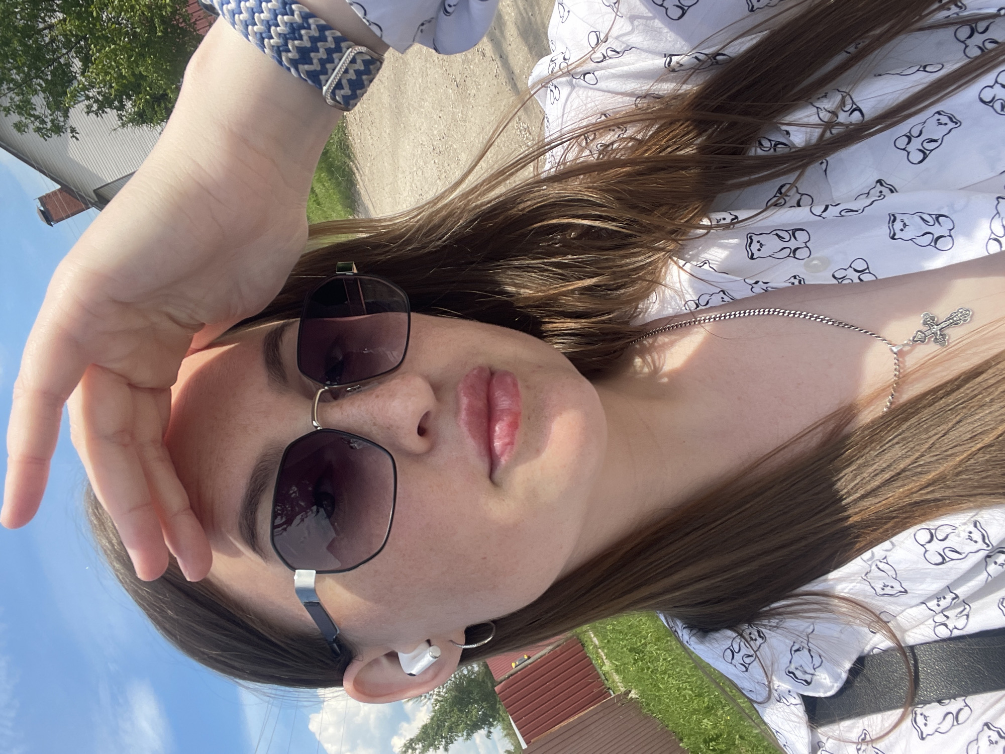 A young woman is captured in a close-up, outdoor selfie-style shot, taken in what appears to be a rural or suburban setting in Сырково, Russia. She is facing the camera, with her right hand raised to shade her eyes from the bright sunlight, which casts shadows across her face.
The woman has fair skin, visible freckles, and light brown or auburn hair that cascades over her shoulders. Her lips appear plump and are slightly parted. She is wearing dark, oversized hexagonal sunglasses with metal accents on the frames. In her left ear, a white wireless earbud is visible, along with a small silver hoop earring. On her right wrist, a wide woven bracelet in blue and white zigzag patterns is worn. She is dressed in a white collared shirt patterned with black outlines of teddy bears. A thin silver chain necklace with a decorative silver cross pendant hangs around her neck.
The background reveals a clear blue sky with some scattered white clouds in the upper left. To the left, a brown slatted fence is partially visible, bordering green grass. To the right, a dirt road or path stretches into the distance, and further back, a white house with a red-brick chimney is visible, partially obscured by green trees under the bright sun. The overall impression is one of a sunny day.