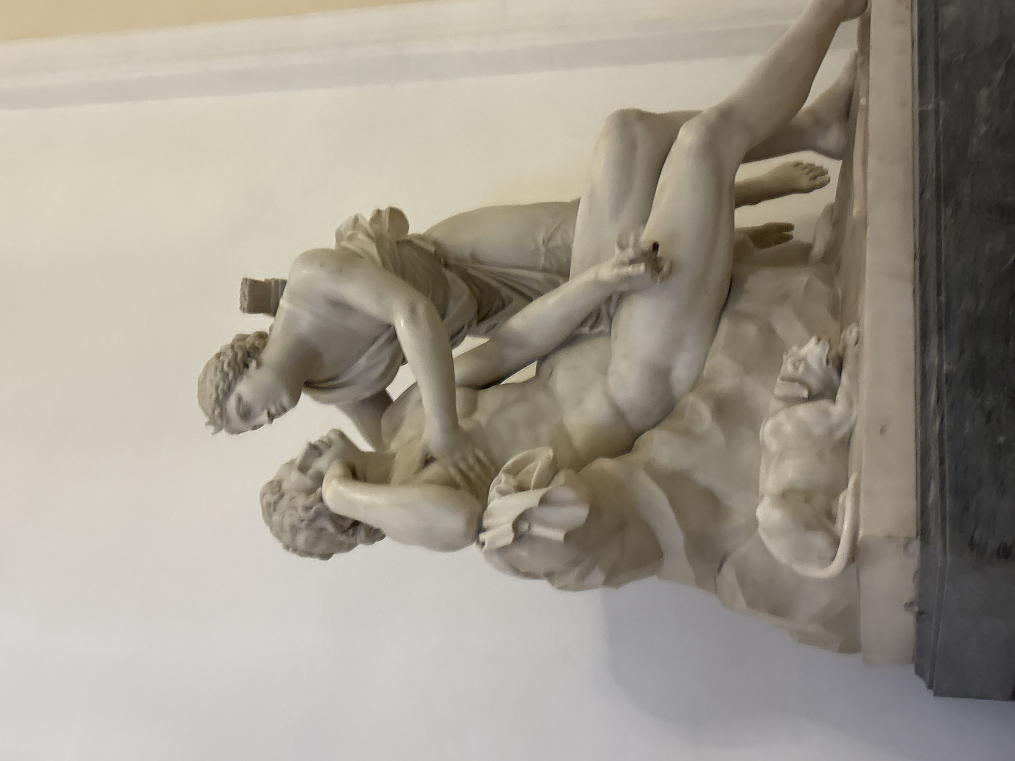 The media file is a photograph of a marble sculpture depicting two figures, likely mythological or allegorical. One figure is lying down, appearing to be struggling or in distress, while the other figure is posed above them, exerting force or in control. The sculpture is intricately detailed, showcasing the musculature and drapery of the figures, carved from light-colored marble. The sculpture is set against a plain, light-colored wall, with a dark vertical element, possibly a frame or pedestal, visible on the right side of the image. The lighting suggests an indoor setting, perhaps a museum or gallery. The overall impression is one of dynamic tension and dramatic action captured in stone. There is no visible text in the image.