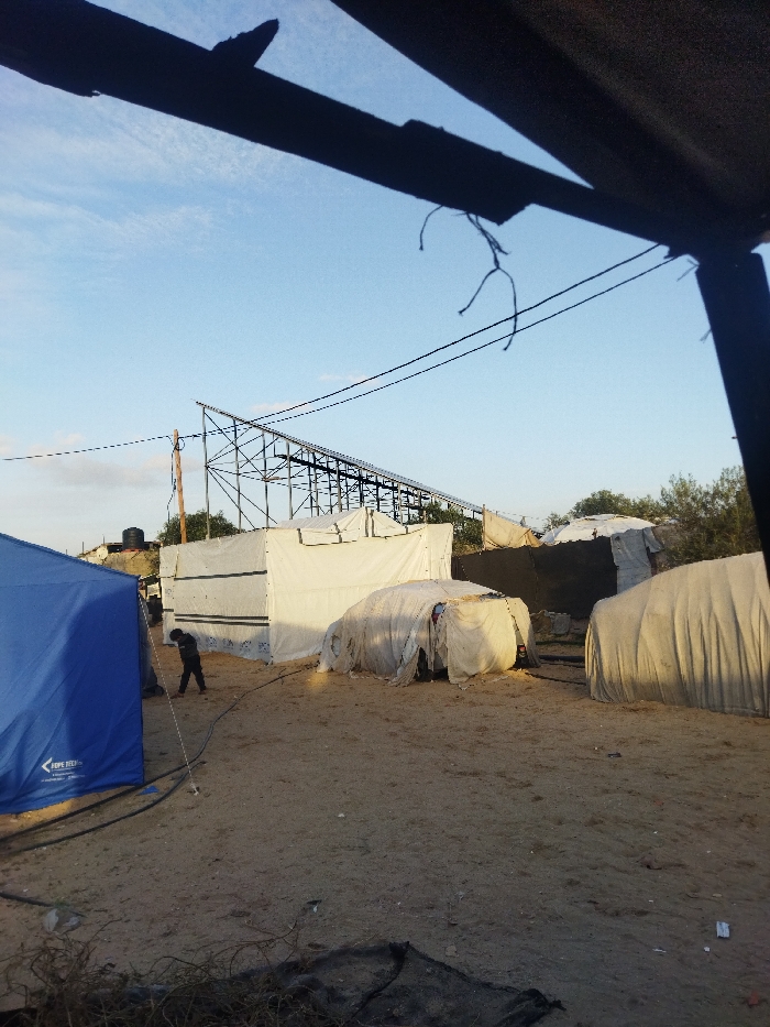 This image presents an informal settlement scene in the city of Khan Yunis, Palestinian Territory, under a clear blue sky with minimal cloud cover.
In the immediate foreground, dark, out-of-focus structural elements, consistent with an overhead covering, are visible at the top and right, from which frayed cords hang. The ground throughout the visible area is primarily packed sand and dirt, with scattered small debris.
To the left in the mid-ground, a partially visible blue tent, marked with "HOPE" near its base, is present. A black hose or cable extends across the ground from near this tent towards the right. Slightly right of the center, a single individual, dressed in dark clothing, stands facing downwards. Behind this person, a rectangular structure constructed from white tarpaulins is erected. To the right of the individual, a vehicle is entirely covered by white tarpaulins. Further to the right, another rounded, tent-like structure, also made of white tarpaulin, is visible.
A prominent metal truss structure, potentially supporting solar panels or a large covered area, spans horizontally across the mid-ground horizon. A wooden utility pole with multiple overhead wires stands to the left of this metal structure, with the wires extending across the upper mid-section of the image. In the distant background on the far left, additional informal shelters and a black water storage tank on a raised platform are discernible. Sparse vegetation, including bushes or small trees, lines portions of the background horizon.