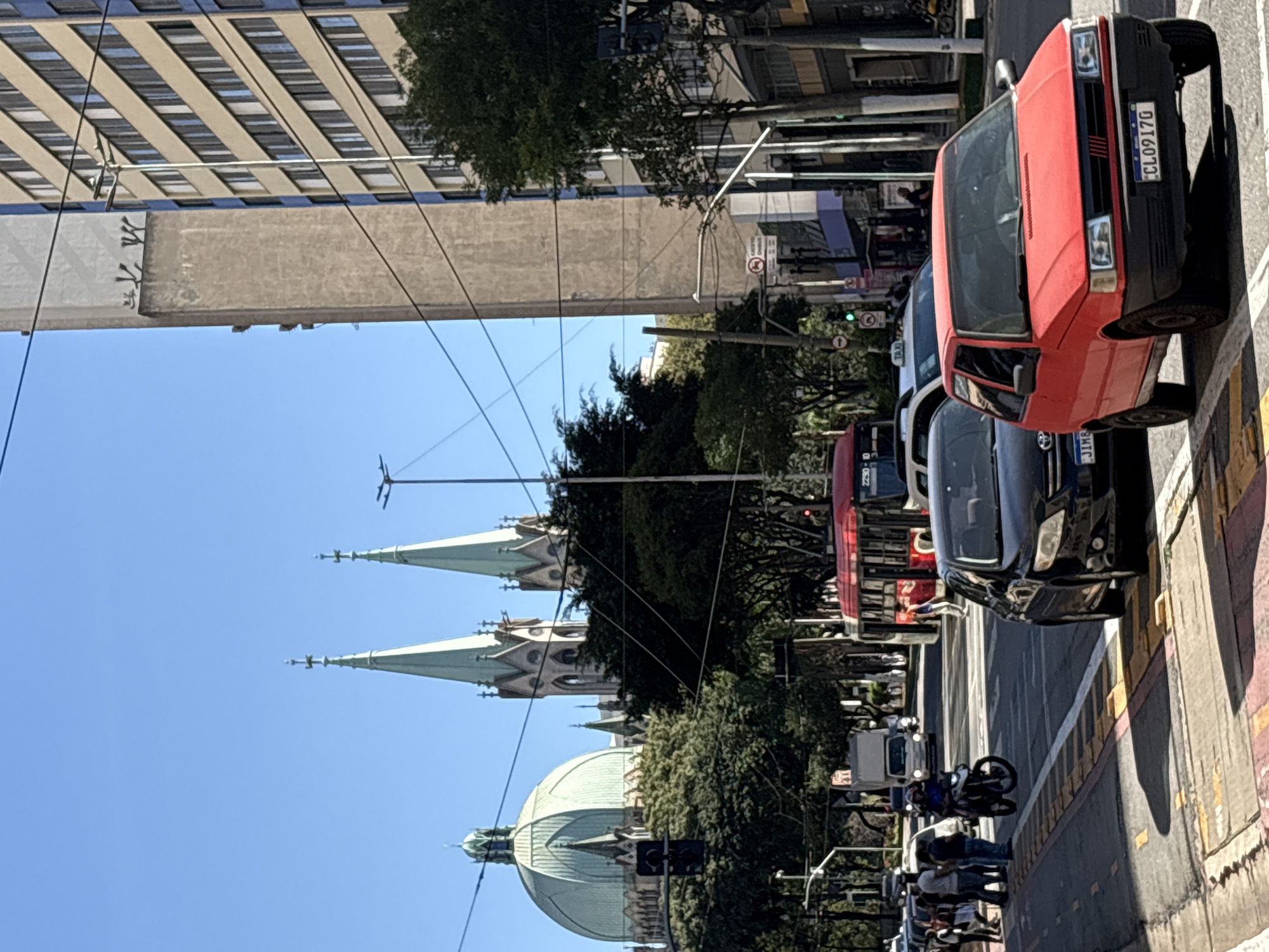 The image captures a street scene in São Paulo, Brazil, with the prominent Metropolitan Cathedral of São Paulo visible in the background. The cathedral features a large green dome and two tall spires. The street is bustling with traffic, including a red bus, several cars, and a motorcycle with a rider. Pedestrians are also present, waiting to cross the street or walking on the sidewalk. The environment appears urban, with tall buildings lining one side of the street and trees providing greenery. The sky is clear and blue, indicating a sunny day. The overall impression is of a lively urban environment with significant architectural and transportation elements.