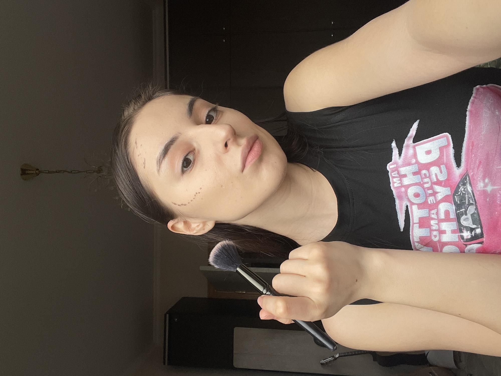 A young woman is posing for a selfie indoors. She holds a black makeup brush in her right hand. Her left arm is also visible. She is wearing a black T-shirt with a pink graphic print that includes the text "MAT." A series of small, dark dots are visible on her face near her ear. A dark wooden cabinet is visible behind her. The scene appears to be lit by artificial light. The location in Moscow, Russia, is not discernible from the image.