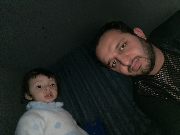 A dimly lit, close-up shot shows a man and a child. The man, with a beard, lies horizontally, head tilted. He wears a dark shirt. To the lower left, a child with dark hair and light skin looks up towards the camera. They wear a light-colored sweater with a ruffled collar. A dark wall with a faint fixture is visible in the background.