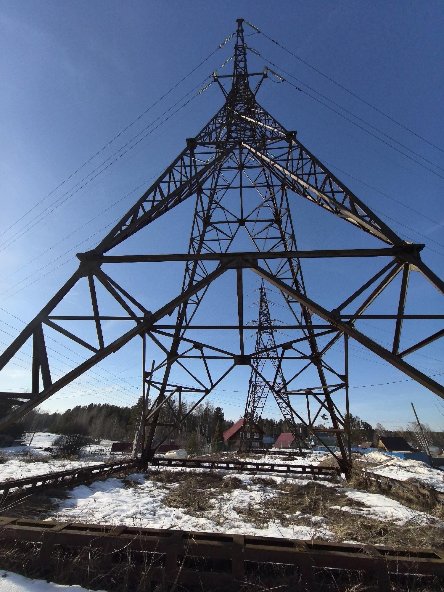 The image depicts two electrical transmission towers in Noviy, Russia. The foreground tower is large, rust-colored, and constructed of steel lattice. It is positioned low to the ground, appearing to be under construction or decommissioned. The ground around it is partially covered with melting snow and patches of dry grass. In the background, visible through the lattice of the foreground tower, is another similar transmission tower, standing upright. Beyond the towers are residential houses with red and dark roofs, interspersed with trees. The sky is clear and blue.