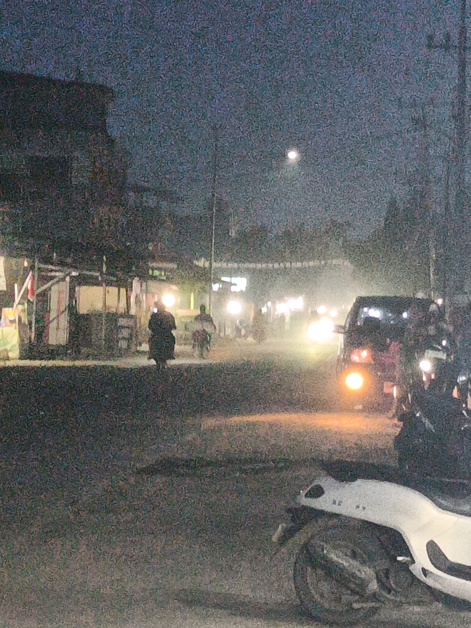 The image depicts a street scene at dusk or night in Sidoarjo, Indonesia, characterized by low light conditions and heavy image noise. A dark, silhouetted building or structure dominates the left side of the frame. Several motorbikes are visible, some with riders. One motorbike in the foreground is predominantly white and appears to be parked or stationary. Further down the street, a dark-colored car is moving, its headlights illuminated, contributing to a hazy atmosphere likely caused by light reflecting off dust or mist. Other vehicles and figures are indistinct in the background due to the low light and image quality. The overall impression is that of a busy street with ongoing activity despite the dim lighting.