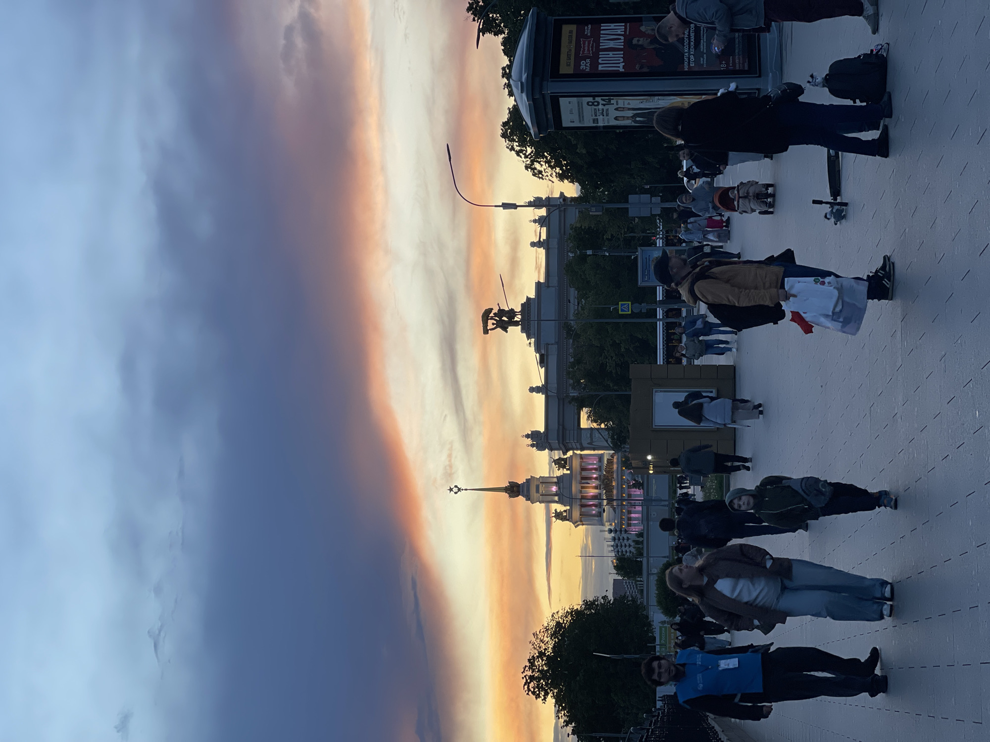 Captured from a ground-level perspective, the image depicts a street scene with pedestrians, set against a backdrop of ornate buildings and a dramatic sunset sky. The sky transitions from a deep, cloudy purple to vibrant hues of orange and yellow, indicating either sunrise or sunset. Silhouetted against the bright sky are detailed building structures, including spires topped with star emblems and classical statues, suggesting a monumental or historical site.
Pedestrians are scattered along a paved walkway, engaging in various activities, such as walking, standing, and interacting with each other. The presence of a billboard with posters indicates an urban setting.
Given the architectural style, including the spires and statues, the location is likely in Moscow, Russia. The architecture resembles the VDNH (Exhibition of Achievements of National Economy) area, known for its Soviet-era monumental structures.