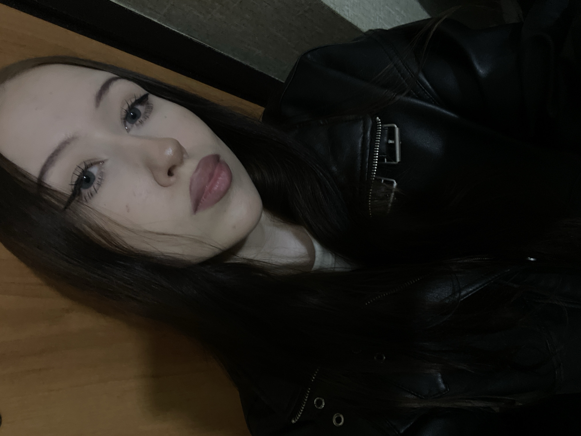 A young woman with long, dark hair and light blue eyes is looking into the camera. She is wearing a black leather jacket over a white t-shirt. Her makeup includes eyeliner and mascara. Her lips are painted with a pinkish-nude lipstick. The background appears to be an interior space with wooden and textured walls. The lighting is dim, creating shadows. The scene is a close-up portrait. The location is not specified in Votkinsk, Russia.