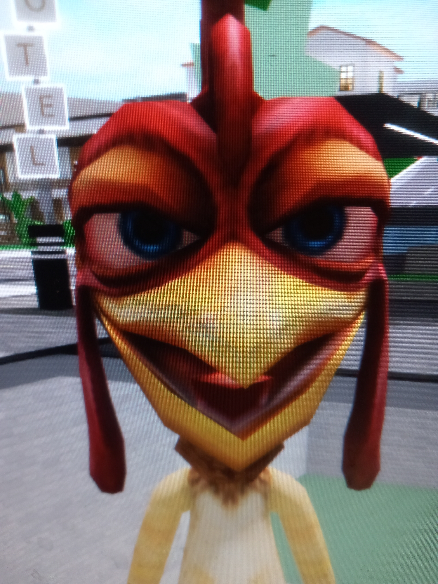 The image is a close-up, slightly pixelated photograph of a cartoonish chicken character. The chicken has a red head with yellow accents, large blue eyes, and a prominent beak. Its body appears to be light yellow or beige. The background suggests an outdoor, urban environment, possibly a game simulation, with buildings and a road visible. There are also some white squares with letters (U, T, E, L) suspended in the air to the left of the chicken. The environment does not provide any specific clues to indicate the location in the city of Rudny, Kazakhstan.