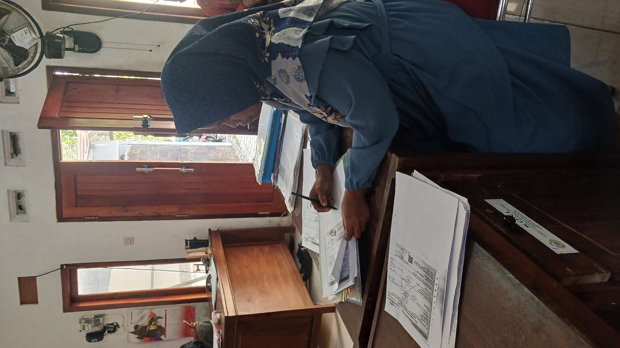 A person wearing a blue hijab and a matching blue dress is sitting at a wooden desk, writing on a stack of papers. The desk is cluttered with documents, folders, and a pen. The environment appears to be an office or administrative space, with wooden doors and windows in the background. A fan is mounted on the wall above the desk. There is a calendar visible on the wall to the left, and a piece of paper with text in what appears to be Indonesian is affixed to the front of the desk. This scene is likely taking place in the city of Kepil, Indonesia.