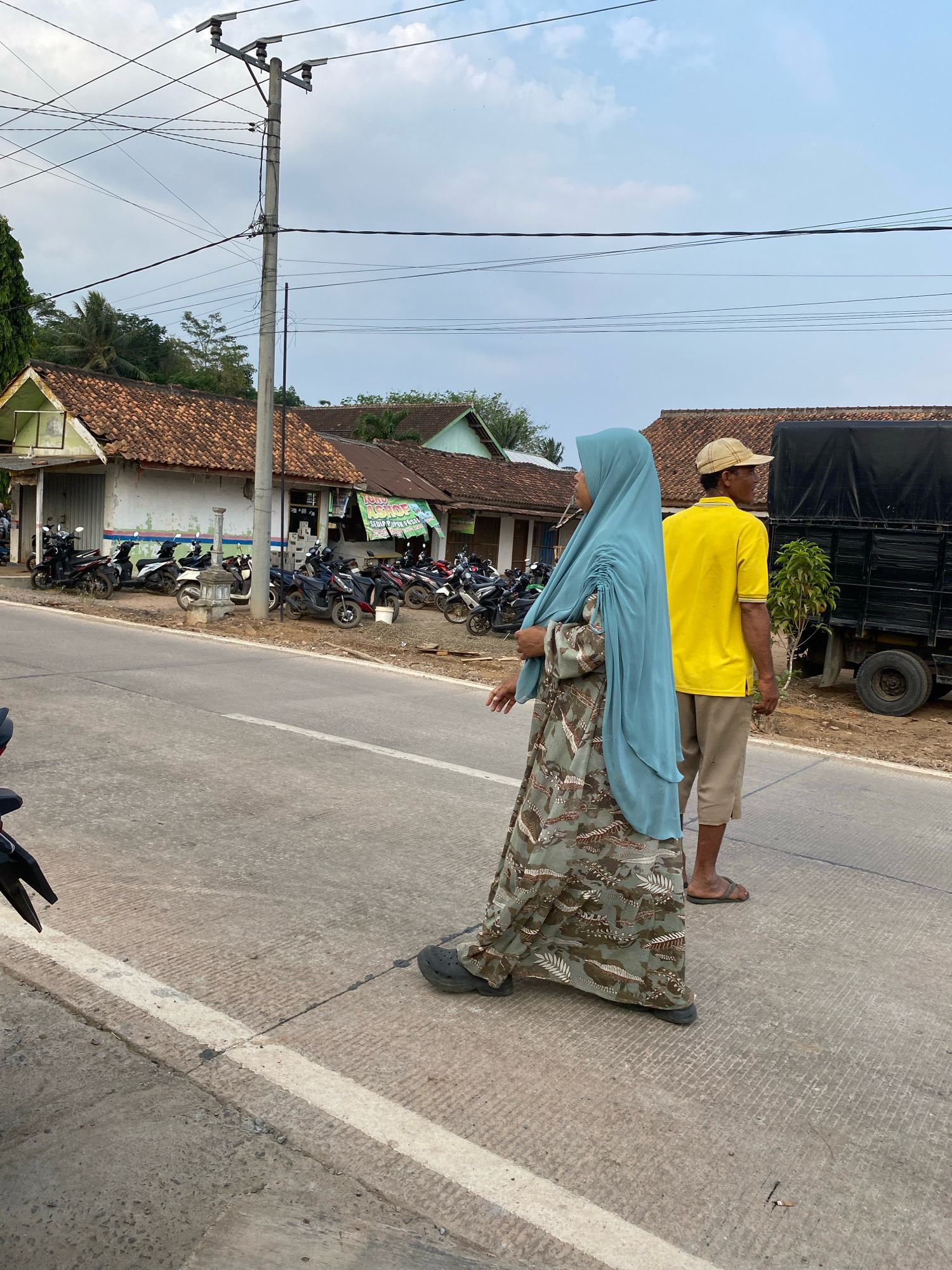 The image depicts a street scene in Sri Sawahan, Indonesia. In the foreground, a woman wearing a light blue hijab and a long, patterned dress walks on a paved road. Beside her and slightly behind, a man in a yellow t-shirt and light brown shorts walks, wearing a tan baseball cap. The road has a white line marking its edge. In the background, several parked motorcycles are visible in front of buildings with red-tiled roofs. A utility pole with numerous wires stands prominently on the left side of the frame. A large black truck is parked on the right side of the road, behind the man. The sky is overcast with some clouds.