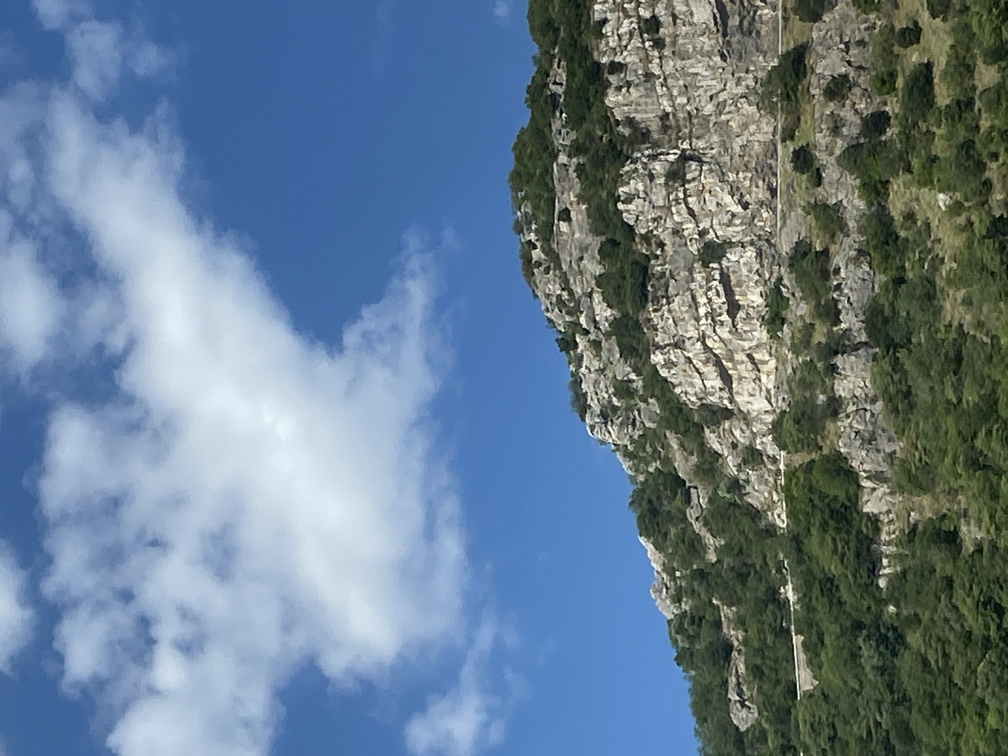 The image displays a view of a rugged, rocky mountainside covered with lush green vegetation, possibly trees and shrubs. A winding road is visible traversing the mountainside. The sky above is a clear, vibrant blue, with scattered white clouds. The terrain suggests a natural, possibly mountainous landscape. This scene is consistent with the coastal mountain ranges found near Budva, Montenegro, known for their dramatic cliffs and verdant slopes overlooking the Adriatic Sea.