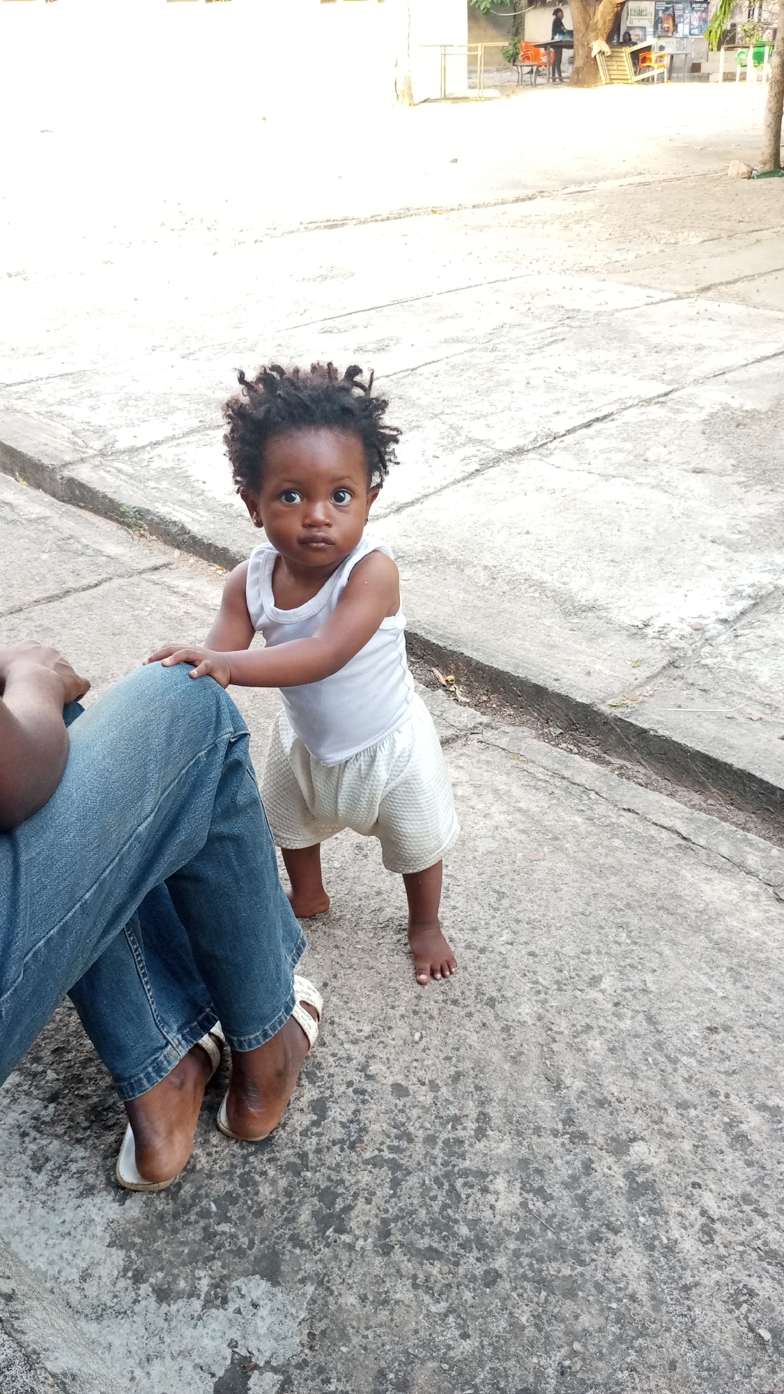 A young child stands on a concrete surface in what appears to be an outdoor area in Adankolo, Nigeria. The child, with dark hair and eyes, is wearing a white tank top and light-colored shorts. The child is holding onto the leg of a person wearing blue jeans and sandals. In the background are trees and what could be outdoor seating. The concrete surface has visible cracks and lines.