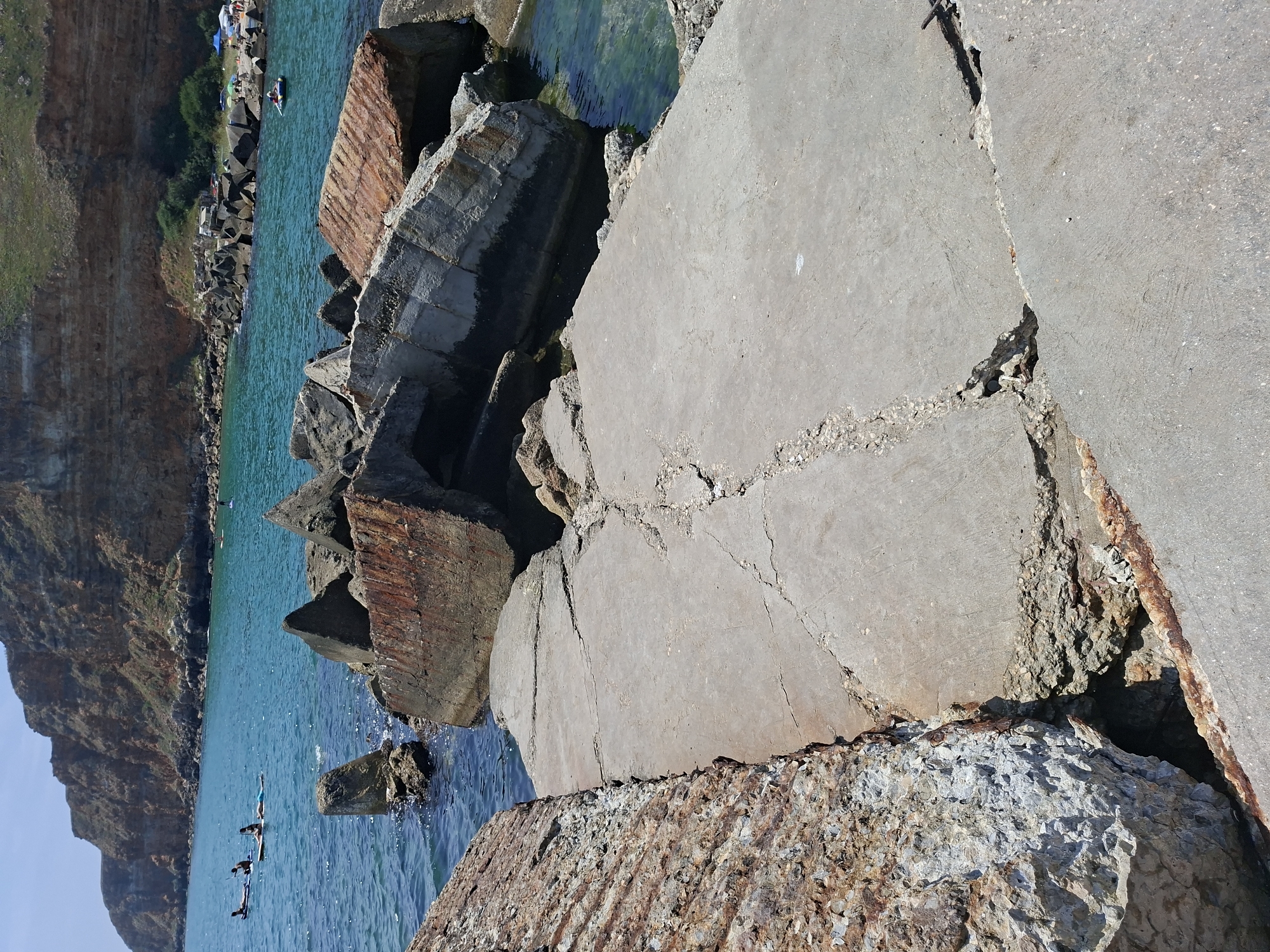 The image depicts a section of the rocky coastline in Balgarevo, Bulgaria, characterized by the presence of large, weathered concrete structures and natural rock formations. In the foreground, a cracked concrete platform is visible, leading towards the sea. The platform appears to be part of an old, possibly abandoned, coastal defense or infrastructure. Beyond the platform, several large, broken concrete blocks, some with rusted metal reinforcements, are scattered in the water and along the shoreline, suggesting a history of erosion and decay.
The sea is a vibrant turquoise color, with clear water allowing a view of submerged rocks and debris. In the middle ground, a few individuals are seen paddleboarding on the calm water, enjoying the coastal environment. Further in the distance, a steep, reddish-brown cliff face rises above the water, covered with sparse green vegetation. The top of the cliff appears to be a plateau with some trees and possibly a beach area with umbrellas and tents, indicating a populated recreational area. The sky is a clear blue, indicating fair weather. The overall scene combines elements of a natural coastal landscape with remnants of human construction, creating a somewhat rugged and historical atmosphere.