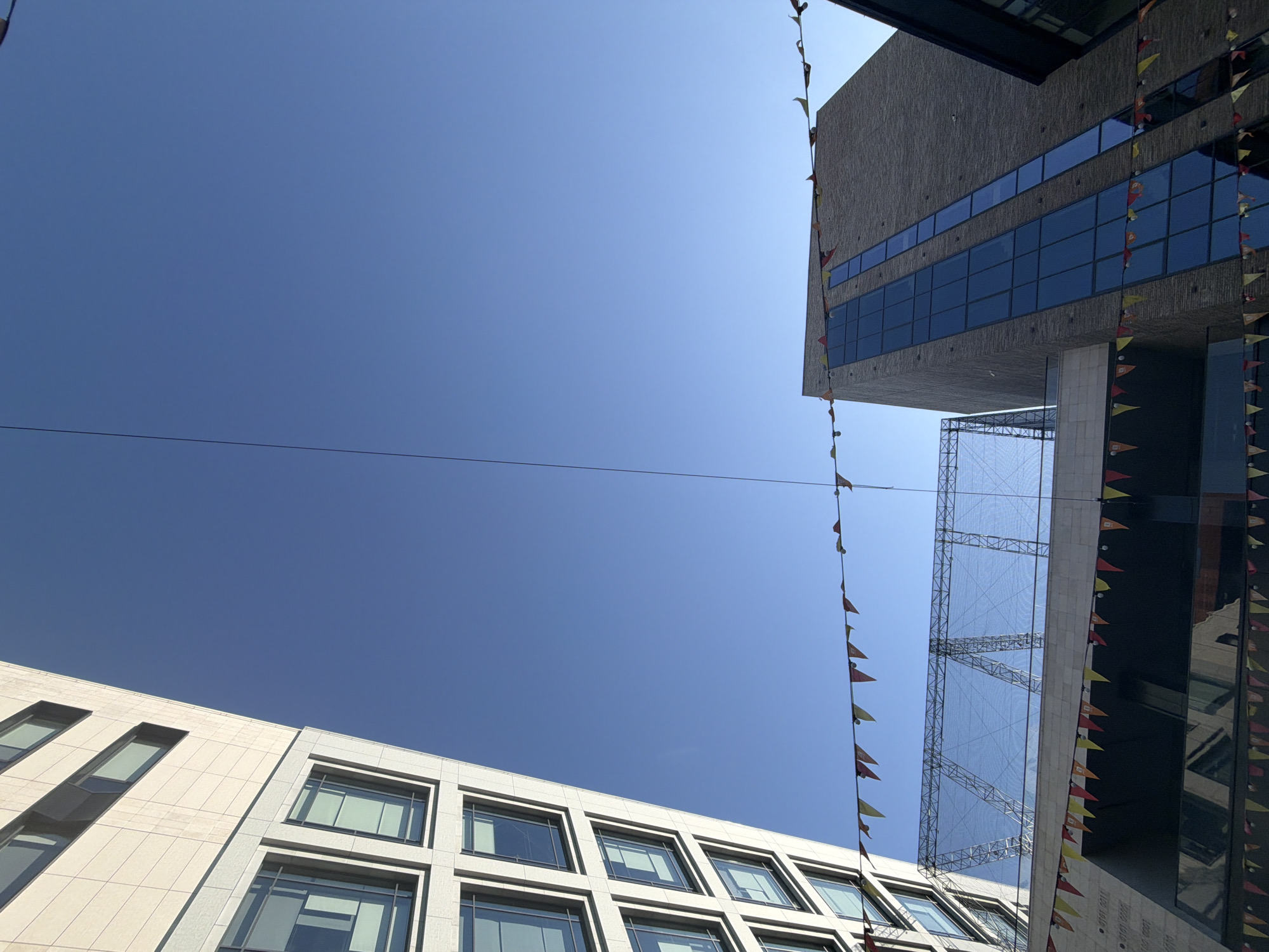 The image captures a low-angle view of buildings in Kazan, Russia, against a bright blue sky. Two contemporary buildings with rectangular windows dominate the frame. The building to the right is constructed from concrete and features a large protruding section with multiple rows of windows. Scaffolding partially obscures this building. The building on the bottom left is a light-colored modern structure. Both buildings are adorned with strings of small triangular flags, suggestive of an outdoor event or celebration. A thin wire stretches across the image.