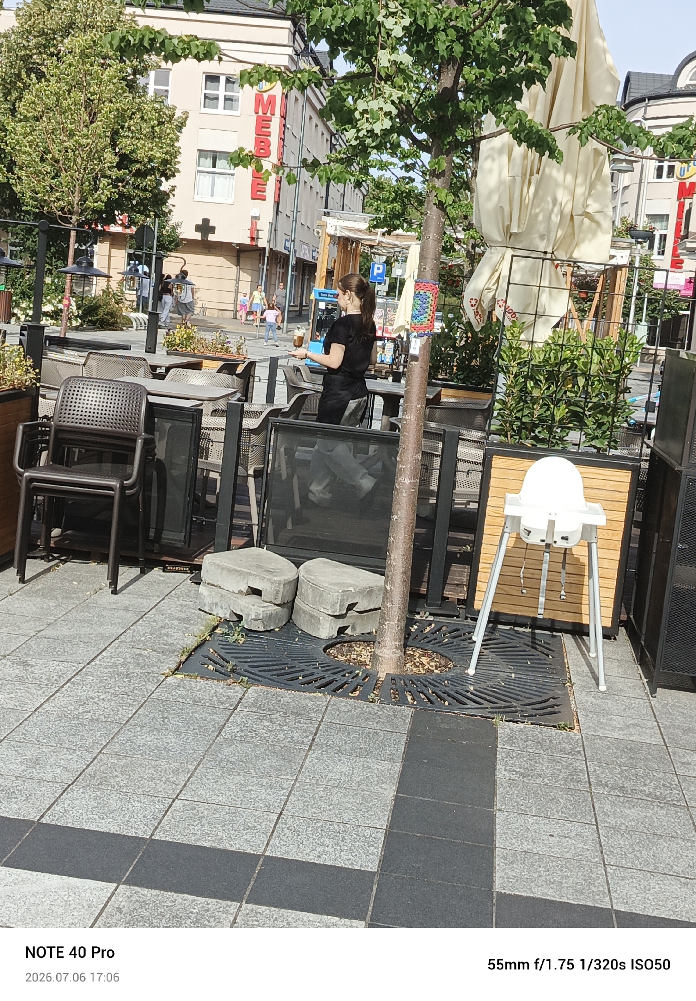 The image depicts an outdoor seating area of a restaurant or cafe on a sunny day in Piaseczno, Poland. In the foreground, several dark brown plastic chairs are stacked, with a metal fence and some grey paving stones. To the right, a white high chair is set up. Behind these elements, a tree with green leaves stands centrally. Further back, there are tables and chairs, and large, folded white umbrellas. A woman in a black shirt is visible, holding a drink. In the background, a building with a sign displaying red letters, possibly for "MEBLE" (furniture in Polish), is seen. The street pavement is made of grey tiles. The date stamp indicates the image was taken on July 6, 2026, at 5:06 PM.