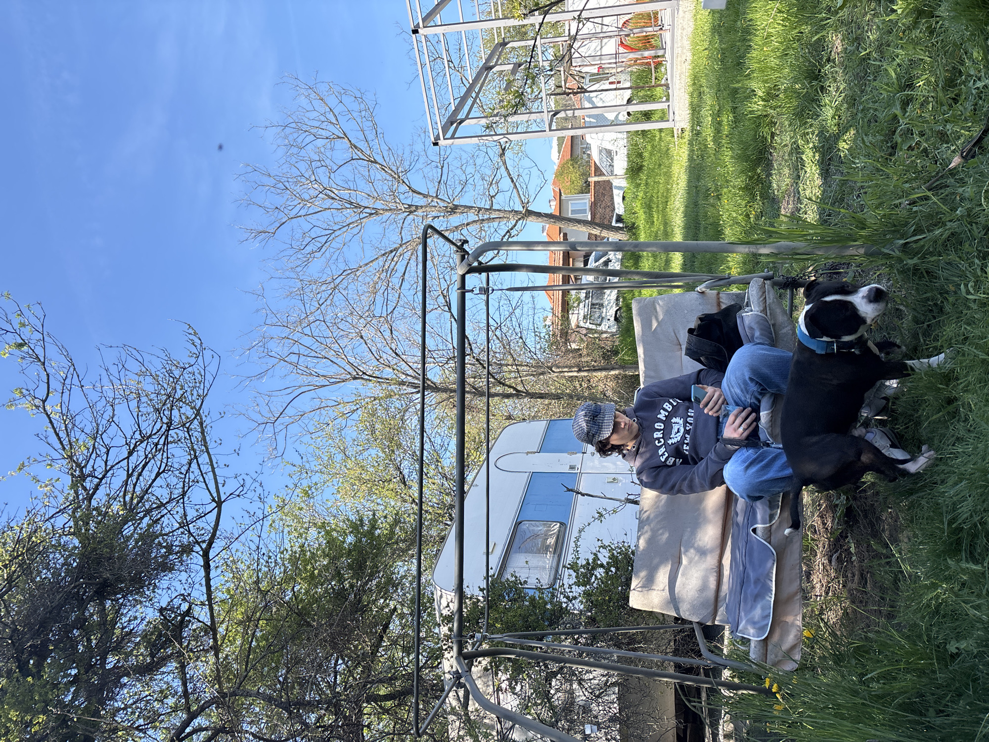 A person sits on a gray cushioned swing in a yard under a clear blue sky. The individual wears a gray hooded sweatshirt with "ABERCROMBIE" written on it, jeans, a patterned cap, and holds a phone. A black and white dog with a blue collar is lying next to the person in the grass. The swing is metal, and behind it, there is a white and blue camper and trees with sparse foliage. Metal framing structures are visible in the background, with green grass. The location is Verinsko, Bulgaria, as that was provided in the prompt.