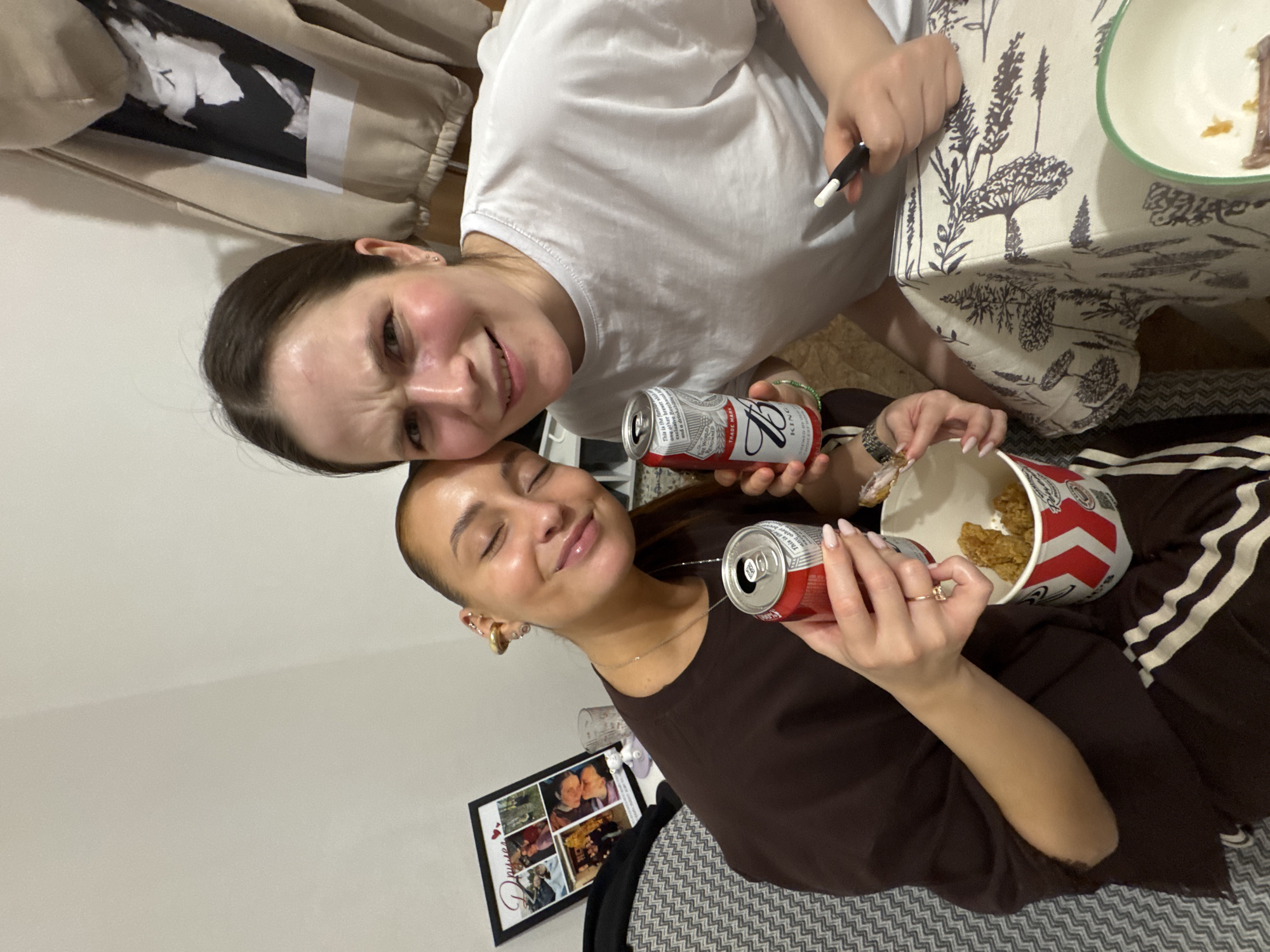 Two women are indoors, the first in a white t-shirt, the second in a brown t-shirt and brown sweatpants with white stripes. They are holding and eating Kentucky Fried Chicken, drinking Budweiser. There is a photo frame on the floor, and a patterned tablecloth. The image was captured in Chaikovsky, Russia.