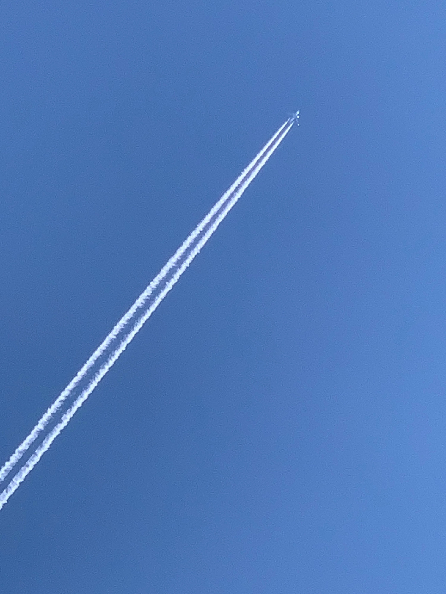 An airplane is flying at a high altitude, leaving a long, white contrail against a clear blue sky. The airplane itself is a small, dark silhouette at the upper tip of the contrail, making it difficult to discern specific details. The contrail is thin and elongated, suggesting the aircraft is moving away from the viewer. The overall scene is one of vast, open sky, with no other objects or elements visible. The location in Prokopyevsk, Russia, is not discernible from the image itself.