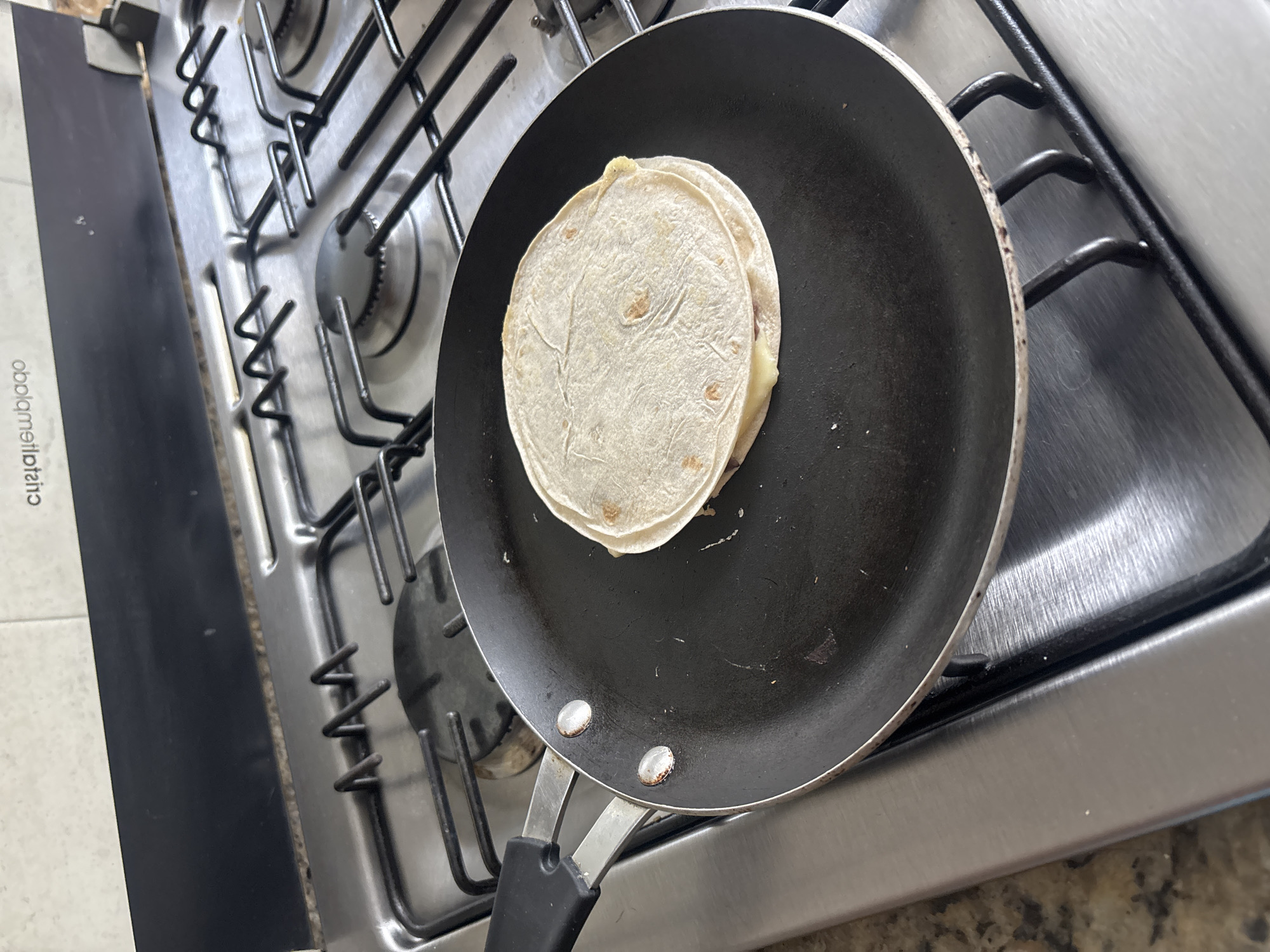 A close-up, angled shot captures a kitchen scene focused on a gas stove. A black non-stick frying pan rests on the metal grates of the stove, which appears to be made of stainless steel. Inside the pan, a folded flour tortilla is being heated, revealing melted cheese oozing from the edges, strongly suggesting a quesadilla in preparation. The pan shows signs of use with some scratches on its surface and slight discoloration around its two rivets. The stove's black metal grates are visible, along with parts of the burners beneath. To the left of the stove, a vertical black panel is visible, on which mirrored text reads "obolameiteiatu". The bottom right of the image shows a speckled countertop. The cooking activity, consistent with preparing a quesadilla, aligns with the provided location context of Puebla, Mexico.