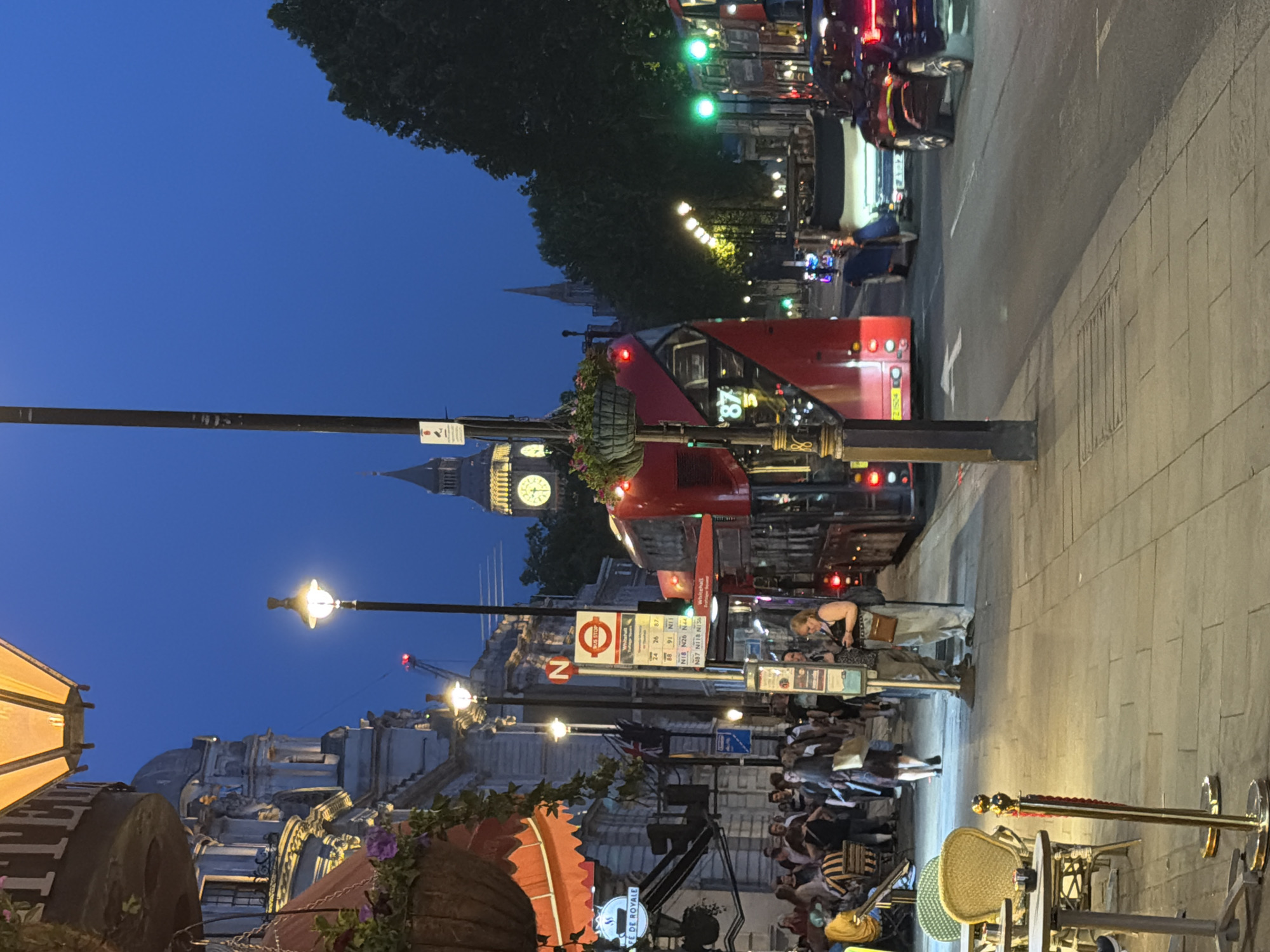 This image depicts a street scene in London, United Kingdom, at dusk or night. The iconic Big Ben clock tower is visible in the background. A red double-decker bus is prominent on the street, suggesting it's a busy route. Streetlights illuminate the scene. People are gathered on the sidewalk, likely waiting for transportation or enjoying the evening. There's outdoor seating for a cafe or restaurant, with chairs and tables visible. A bus stop sign is present, indicating the location as a public transportation hub. The image is slightly tilted.