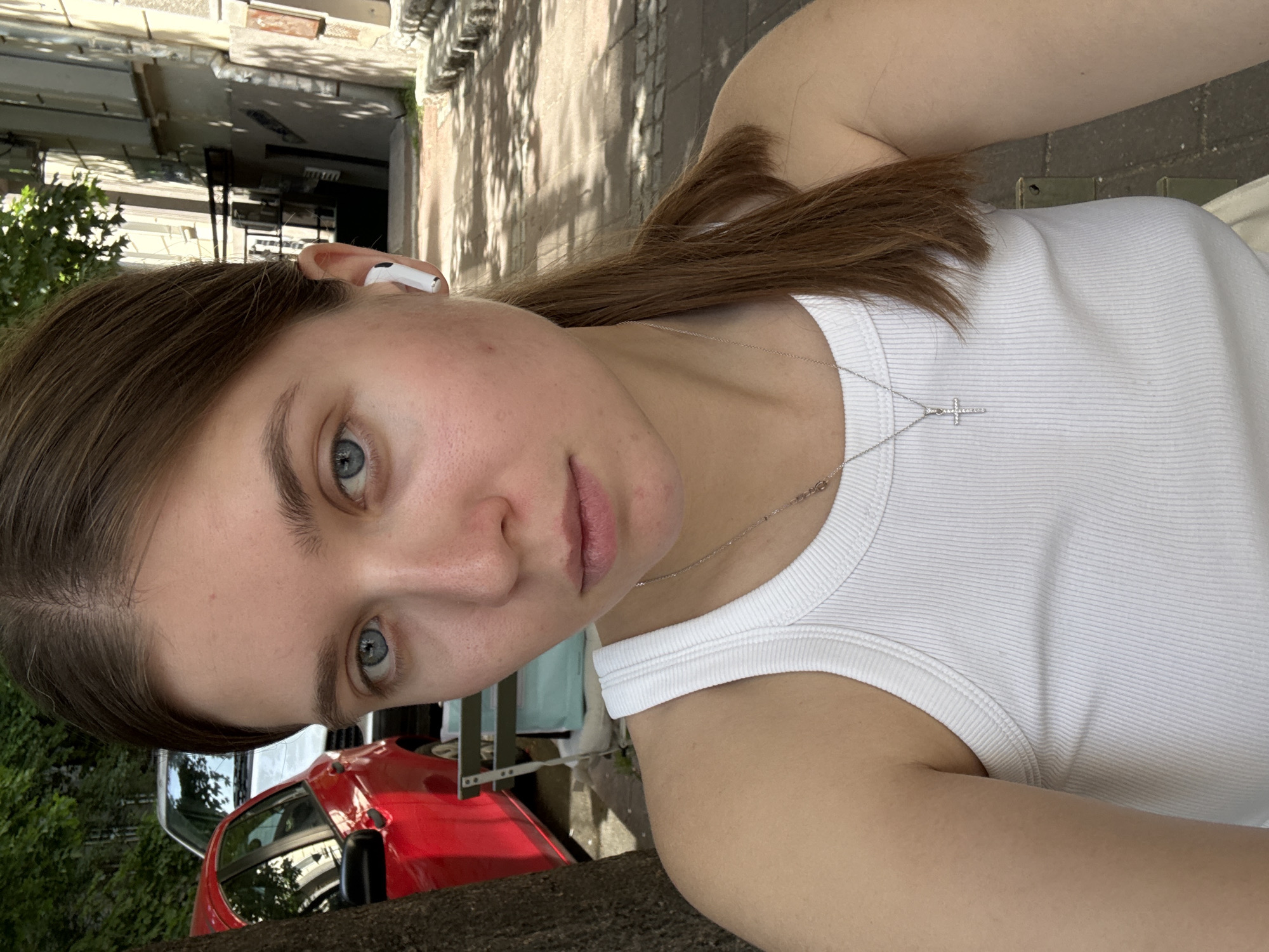 A young woman stands on a street in Belgrade, Serbia, posing for a selfie-style photograph. She wears a white tank top and a silver necklace with a cross pendant. Her light brown hair is pulled back. She has fair skin and blue eyes. An earbud is visible in her left ear. Behind her, a red car is parked on the street. The background shows a building and trees, suggesting an urban residential area with dappled sunlight.