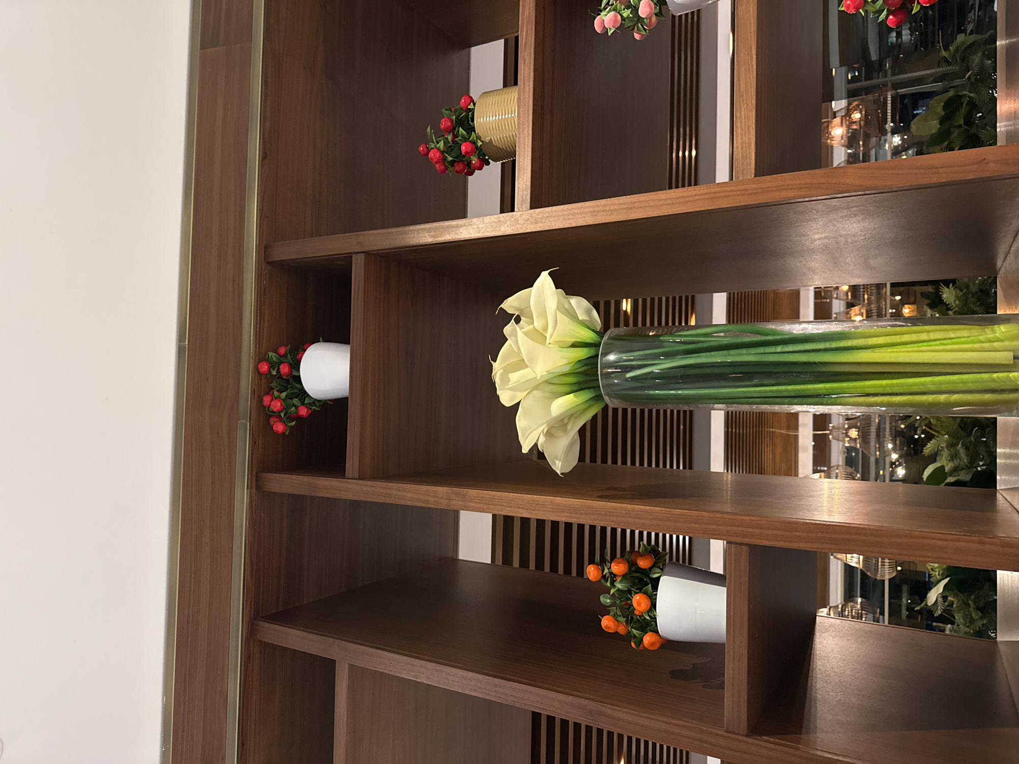 A close-up, low-angle interior shot shows a wooden shelving unit with various decorative items. The shelving unit is made of dark, polished wood and features multiple cubbies. On the upper shelves, there are small potted plants with red berries, one in a white pot and another in a textured tan pot. In the center of the frame, a tall, clear glass vase holds a bouquet of creamy white calla lilies with green stems. The background consists of more shelving, with some out-of-focus glimpses of lights and greenery that suggest a restaurant or hotel setting. The overall impression is one of sophisticated interior design.