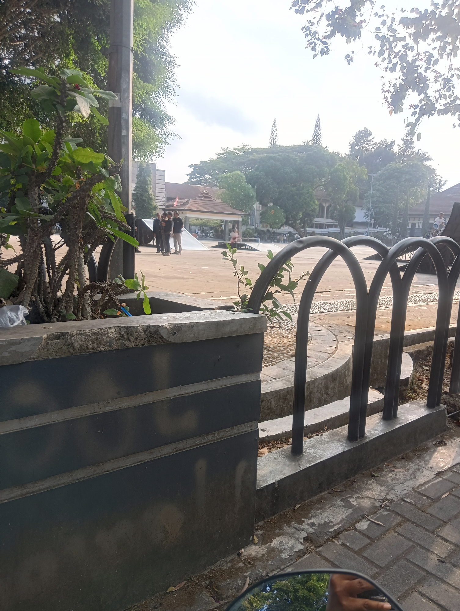 The image shows an outdoor scene in Purwawinangun, Indonesia. In the foreground, a grey concrete planter box with dark grey horizontal slats is visible. A spiky green plant grows from the planter. A black metal fence with arched bars runs along the right side of the planter. Beyond the fence, a paved area with cobblestones is visible. In the background, several people stand in a group. Further back, a building with a brown roof and an open pavilion-like structure are present. Trees and other buildings are scattered throughout the background, and the sky is overcast. A motorcycle mirror reflects a hand and trees in the lower right corner.