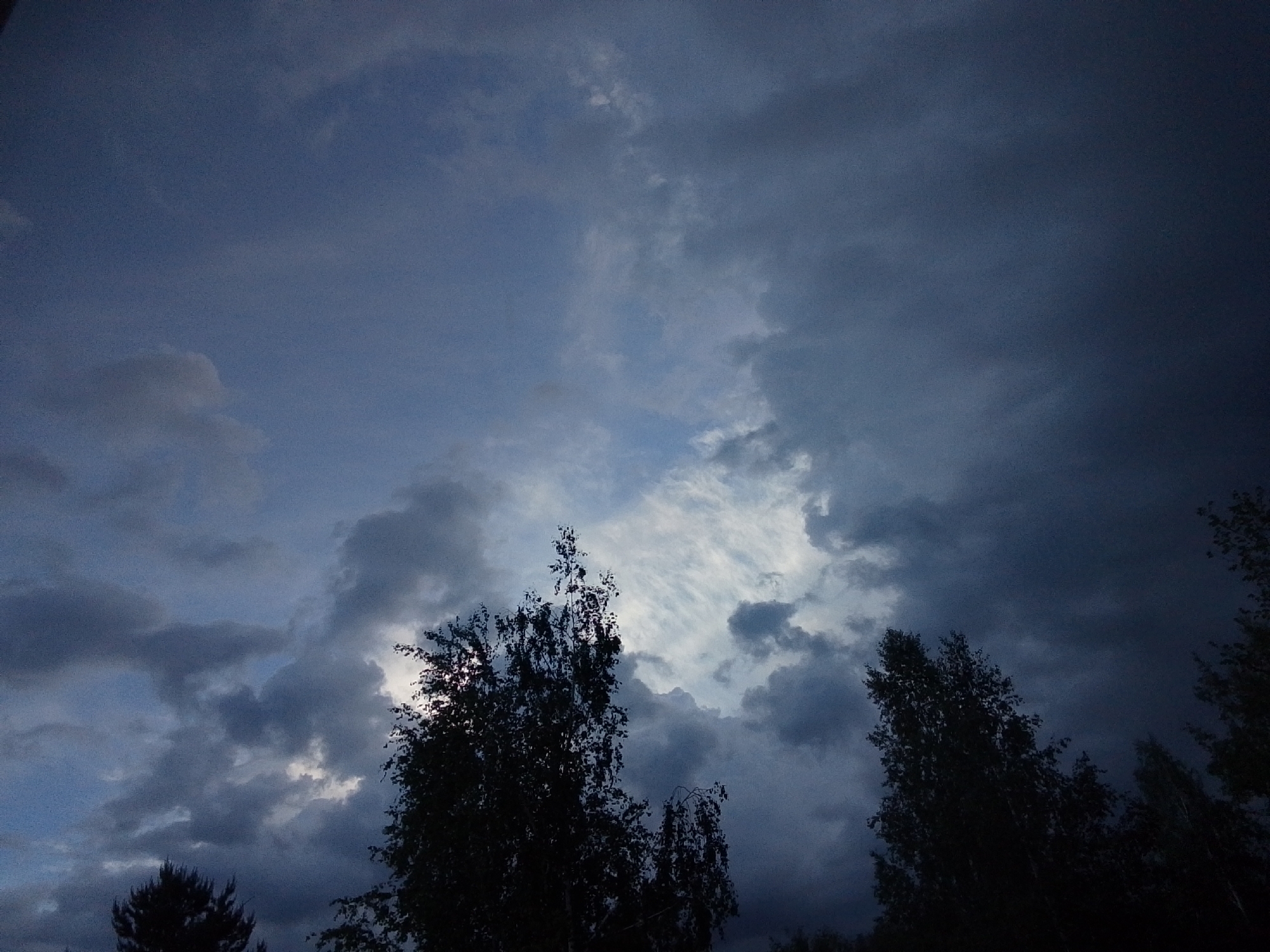 The image shows a low-angle view of a dramatic sky with dark, stormy clouds. The clouds are predominantly gray and blue, with some areas of lighter white clouds visible through gaps in the darker formations. Silhouetted against the sky are several trees, their dark forms contrasting with the lighter sky. The trees appear to be various sizes, suggesting a natural, wooded area. The scene is dimly lit. There is no evidence of any people, subjects, actions or interactions. The location of 1-Go Maya, Russia cannot be proven, only hypothesized.