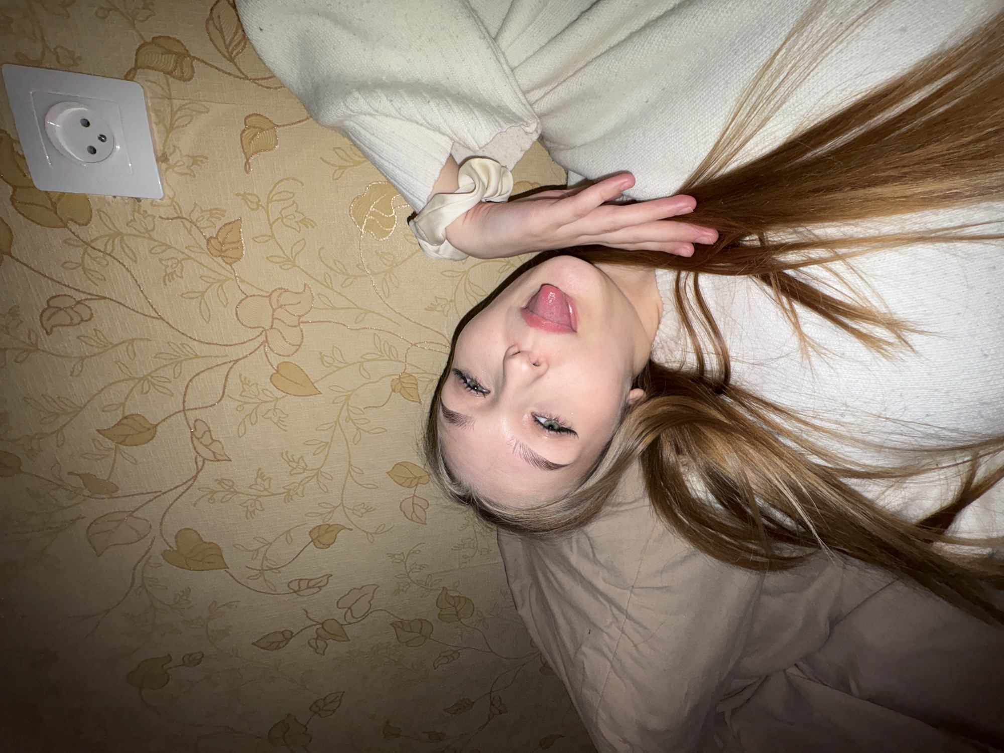 A young woman is captured in a close-up, playful shot, lying down with her head tilted and her tongue sticking out. She has long, light brown hair fanned out, and is wearing a light-colored, possibly cream or white, knitted sweater. A matching scrunchie is visible on her left wrist. Her eyes are open and looking upwards, with prominent eyebrows and eyelashes.
The background consists of a light beige wall covered in wallpaper featuring a delicate pattern of brown and gold leafy and floral designs. In the upper left portion of the image, a white electrical outlet with two circular holes and a ground pin opening (a Type F socket) is clearly visible on the wall. A crumpled light brown or beige blanket or duvet is partially visible underneath her head and extends towards the lower right of the frame, suggesting she is resting on a bed or similar soft surface.
The scene appears to be an indoor setting, likely a bedroom, given the blanket and wallpaper. The presence of a Type F electrical outlet is a specific detail consistent with the provided location context of Chaikovsky, Russia. The overall impression is one of casualness and lightheartedness. No text is visible in the image.