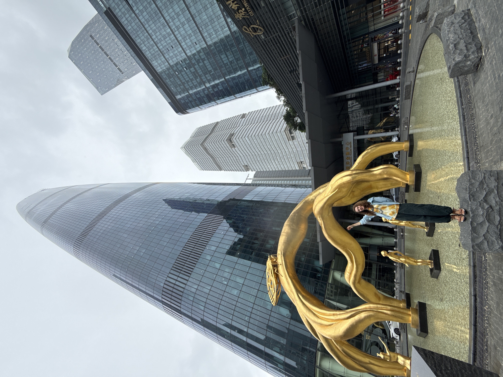 The photo depicts a woman standing in front of a large, golden sculpture in what appears to be an urban environment. The woman is posing with one arm outstretched, wearing a denim jacket, a tie-dye shirt, dark trousers, and sandals. The sculpture behind her is a stylized, abstract piece with what looks like golden ribbons and figures.
In the background, a towering, modern glass skyscraper dominates the scene. Other buildings of varying heights and designs can also be seen. The sky appears overcast, suggesting a cloudy day. The photo appears to have been taken in Tianhe District, China, given the presence of distinctive skyscrapers and urban planning characteristic of this area. The ground in front of the sculpture features a water feature or fountain.