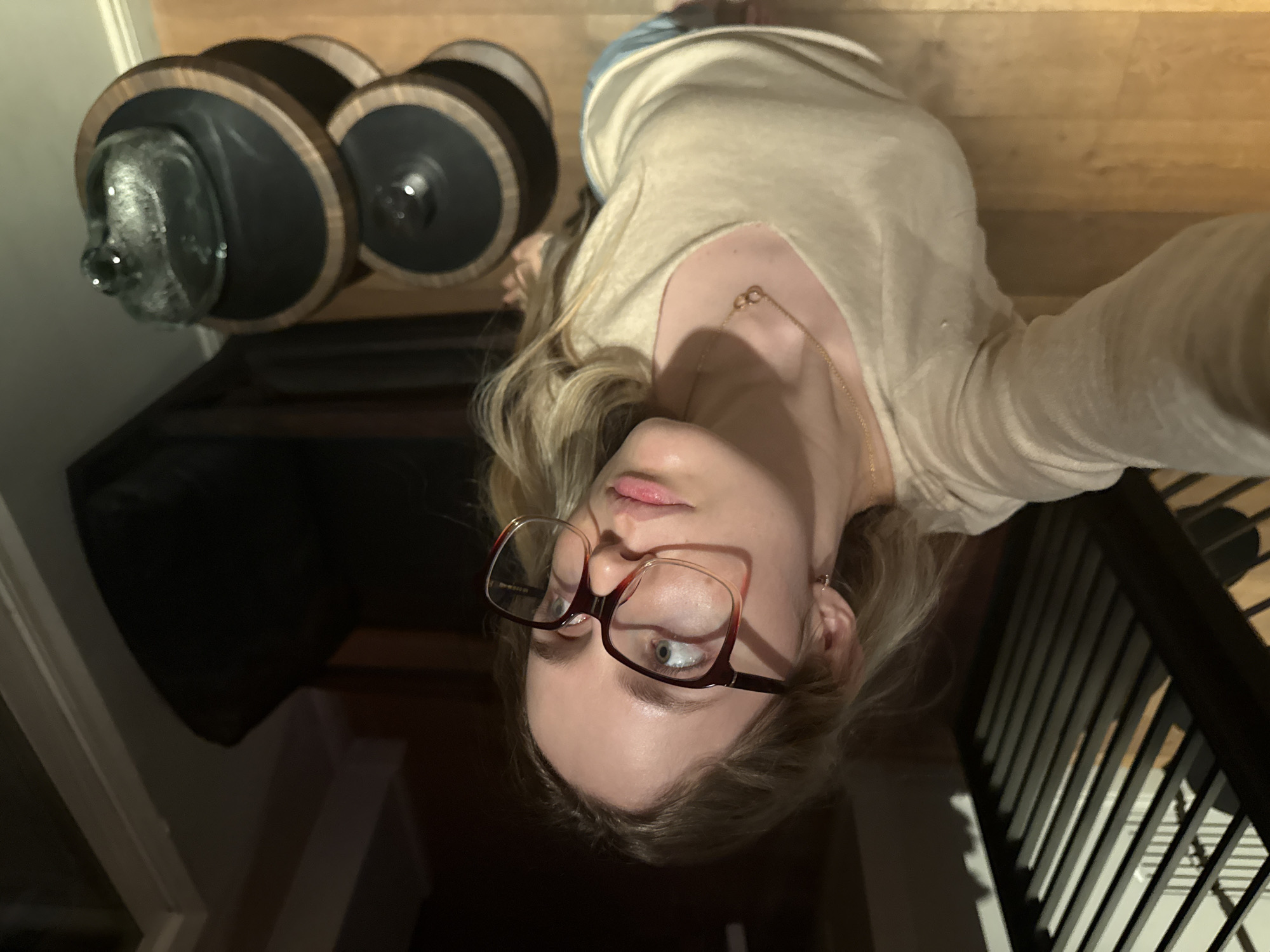 A woman with fair skin and light blonde hair, wearing red-framed glasses, a beige v-neck shirt, and a gold necklace, takes a selfie from a low angle. Her head is tilted to the right. To the left of the frame are three black and wood decorative objects. To the right of the frame is a dark railing. The floor is wood. The location is possibly an indoor space, potentially in Mexico City, Mexico. There is no way to ascertain the precise location within Mexico City.