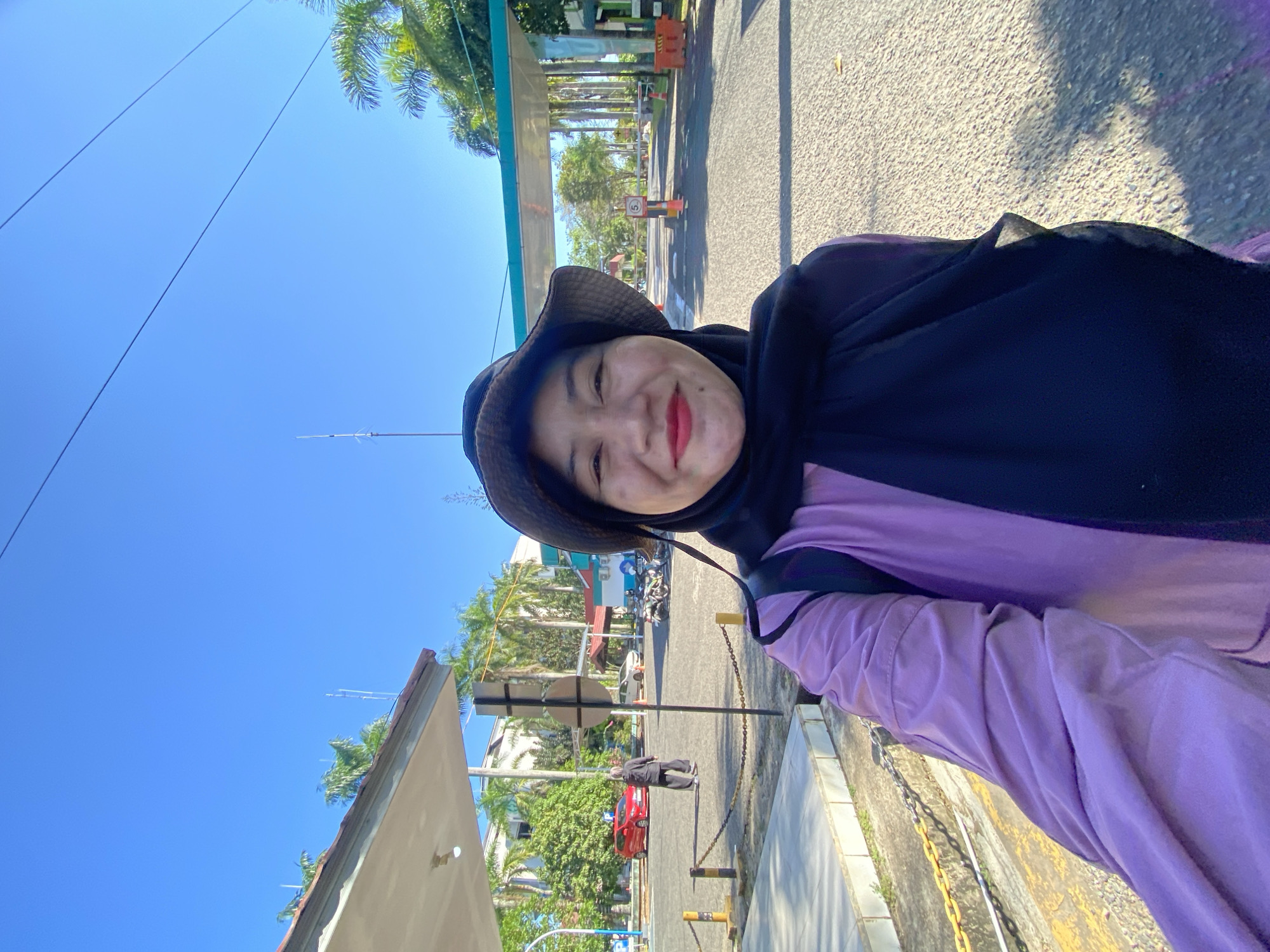 A woman wearing a wide-brimmed hat and a black hijab, with bright red lipstick, smiles at the camera. She is wearing a purple and black patterned tunic. She is standing in what appears to be an outdoor area with a paved road and palm trees visible in the background. The location is identifiable as the entrance gate to Lambung Mangkurat University (UNLAM) Banjarbaru campus in Banjarbaru, Indonesia, characterized by the distinctive campus gate structure and university buildings. A red car is parked to the left, and several motorcycles are parked in the background to the right. The sky is clear and blue.