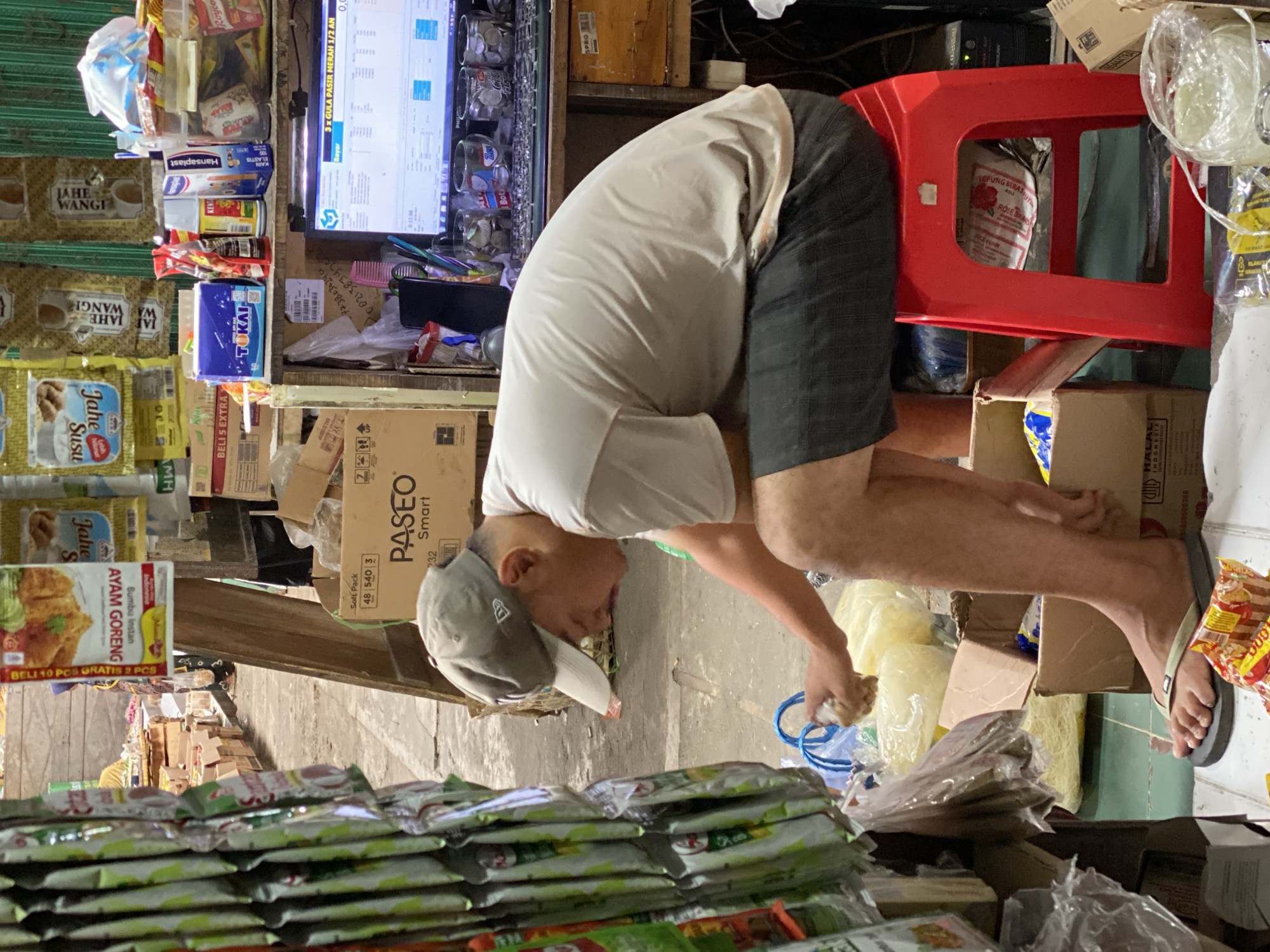 In Ambarketawang, Indonesia, a person wearing a grey baseball cap and a light-colored t-shirt is seated on a red plastic stool in what appears to be a market stall or small shop. The individual is wearing dark shorts and flip-flops. They are hunched over, reaching into a pile of yellow, dry goods that are in plastic bags. In front of them, on the floor, are several packets of food items. Behind the person, a computer monitor displays a point-of-sale or inventory screen. The stall is filled with various products, including packaged food items, boxes, and bags, suggesting a retail environment. The overall setting is cluttered, with goods stacked and displayed throughout the space.