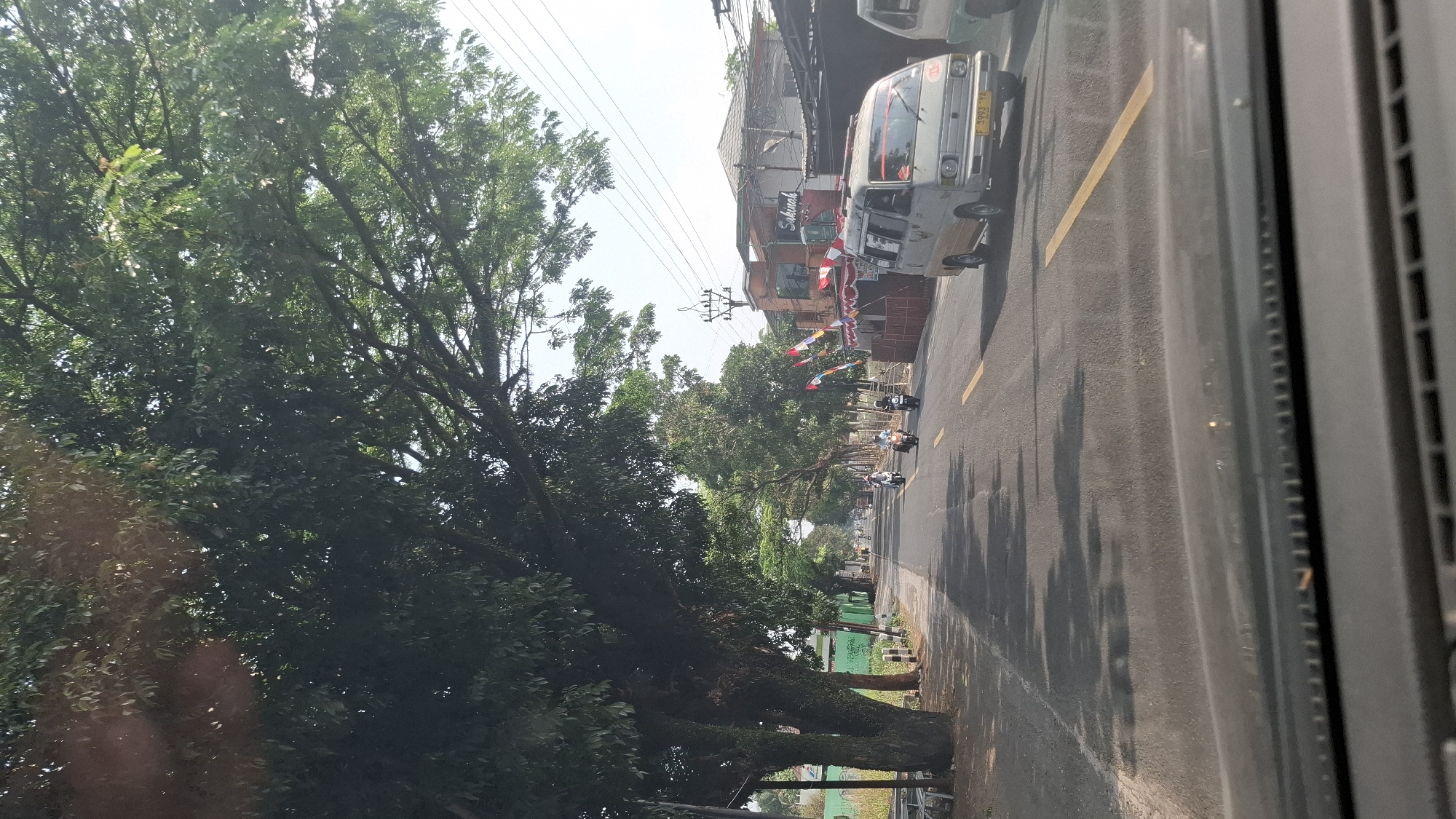 The image depicts a street scene in Cikaroya, Indonesia, under a bright sky with a few wispy clouds. Large, mature trees with dense green foliage line the left side of the road, casting dappled shadows on the asphalt. The road itself is paved and features a prominent yellow dashed line marking the center.
Several motorcycles are in motion on the road, with two riders clearly visible in the middle distance. To the right, a light-colored van is traveling in the same direction. Behind the van, a building with signage that includes the word "Sakuna" is visible, adorned with red and white striped banners. Further buildings and utility poles are present in the background, suggesting an urban or semi-urban environment. The perspective is from within a vehicle, looking forward through the windshield, which has some reflections on its surface. The foreground shows the dashboard of the vehicle.