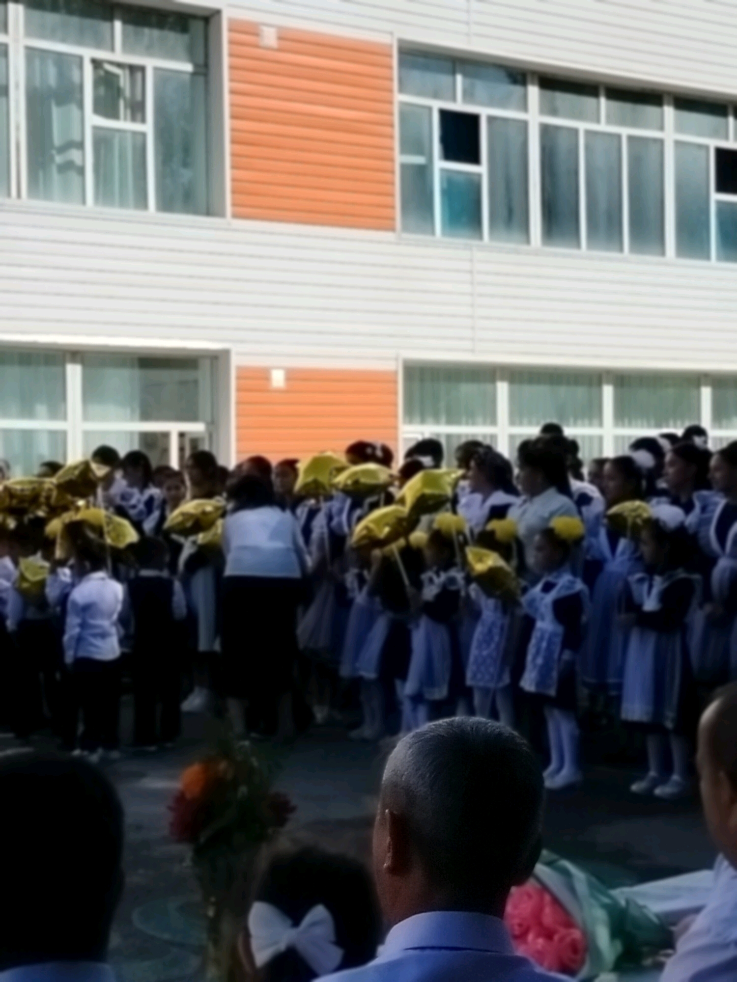 The image depicts a group of children, likely students, gathered in front of a building. Many of the children are holding yellow balloons, and some appear to be wearing school uniforms. The building has a modern architectural style with large windows and horizontal siding. The ground in front of the building is paved. In the foreground, the backs of two adult men's heads are visible. The overall scene suggests an outdoor event, possibly a school celebration or ceremony. The location is identified as the city of Kölkent, Kazakhstan.