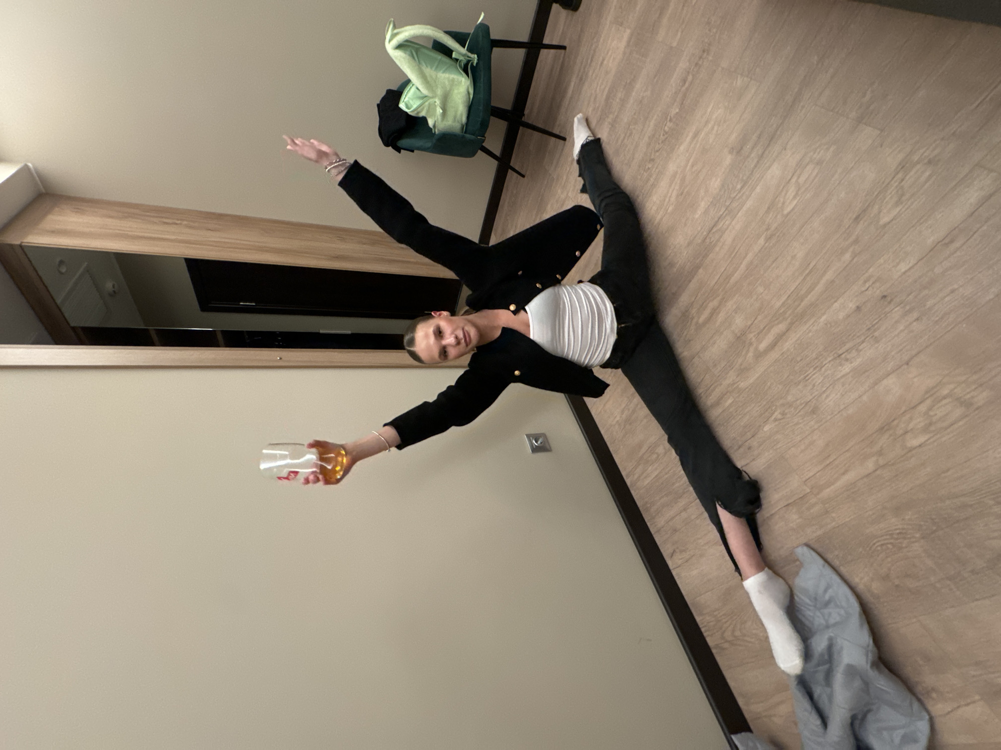 A young Caucasian woman is performing a split on a wooden floor, holding a glass of amber liquid in her right hand, raised above her head. She is wearing a white tank top, a black blazer with gold buttons, and black jeans that are ripped at the knees. Her left foot is extended forward, and her right leg is extended backward. She has white socks on both feet. Her arms are outstretched, with her right arm raised and her left arm extended to the side. She is looking at the camera with a neutral expression.
The background shows a plain beige wall with a silver electrical outlet. To the right, there is a dark green armchair with a light green backpack draped over it. Further to the right, a tall wooden closet with mirrored doors is visible. The lighting appears to be artificial.
The scene does not provide any explicit indicators to pinpoint its location within the city of Saint Petersburg, Russia. The depicted environment is generic and could be found in various urban settings.