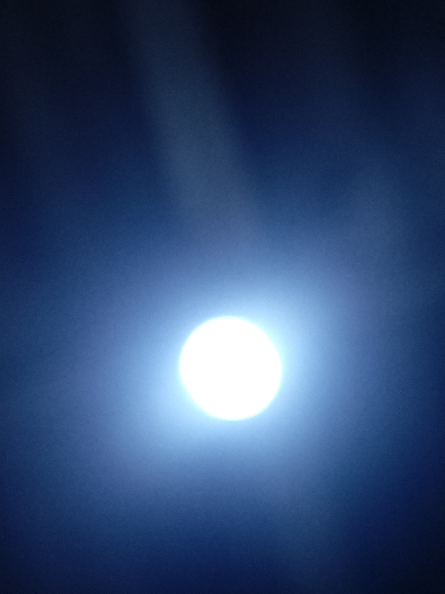 The image shows a bright, circular light source against a dark blue background. The light source dominates the center of the frame. The surrounding area is dark blue, with subtle variations in tone, possibly due to atmospheric conditions or the quality of the image capture. There are no visible people, subjects, or objects besides the light source and the blue background. The image suggests it could be a night scene with a strong light source, possibly the moon. There is no definitive indication of the specific location of Juan Griego, Venezuela in this image.