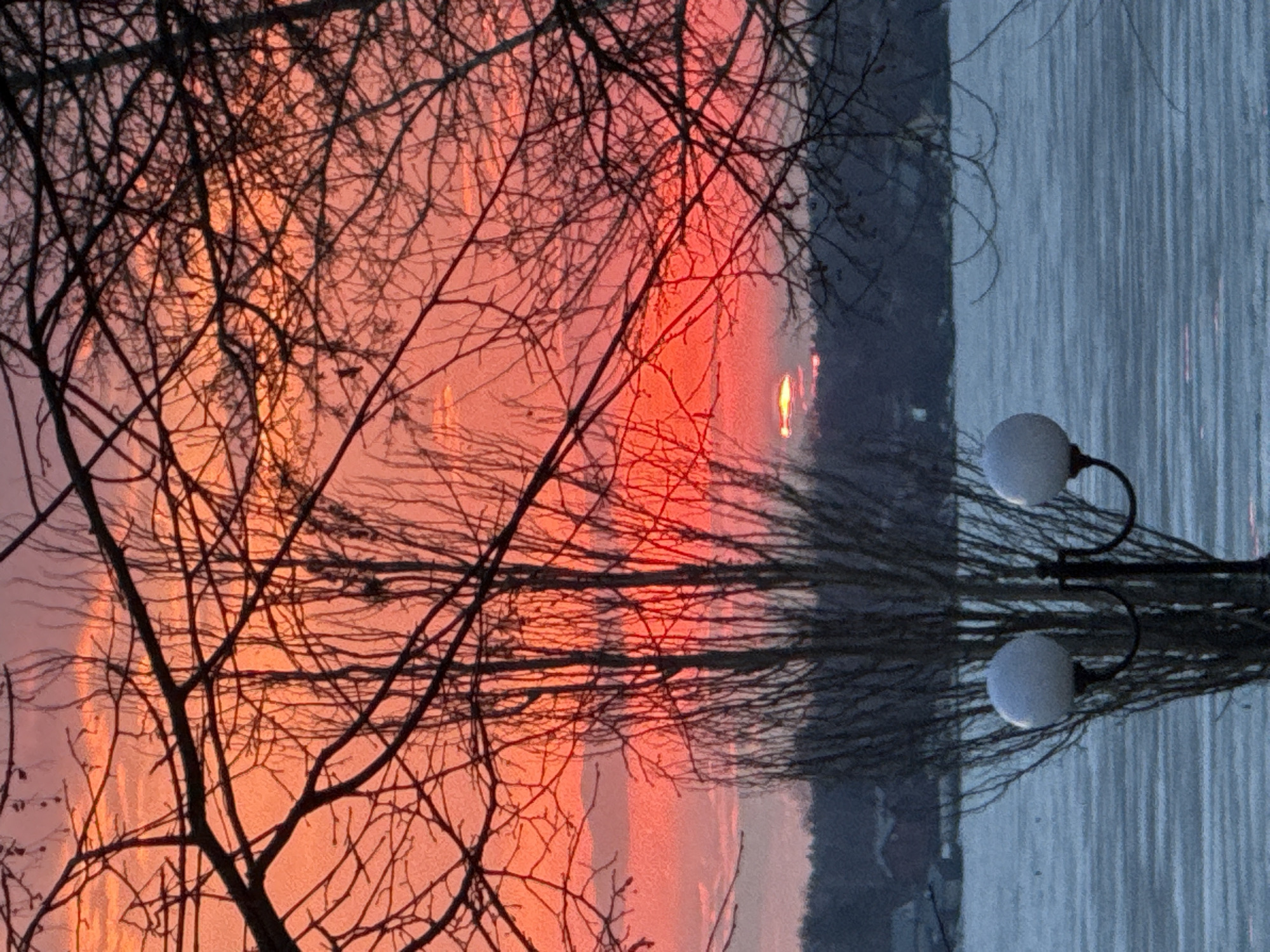 The image presents a view of a body of water, likely a lake or reservoir, at sunset or sunrise, possibly in Votkinsk, Russia. The sky displays vibrant hues of orange and red, indicative of the sun's position near the horizon. Silhouetted tree branches are prominent in the foreground, adding depth and texture to the scene. On the right side of the image, a lamppost with a spherical light fixture is visible. The water reflects the colors of the sky and the light from the lamppost, enhancing the overall atmospheric effect. The composition suggests a tranquil environment.