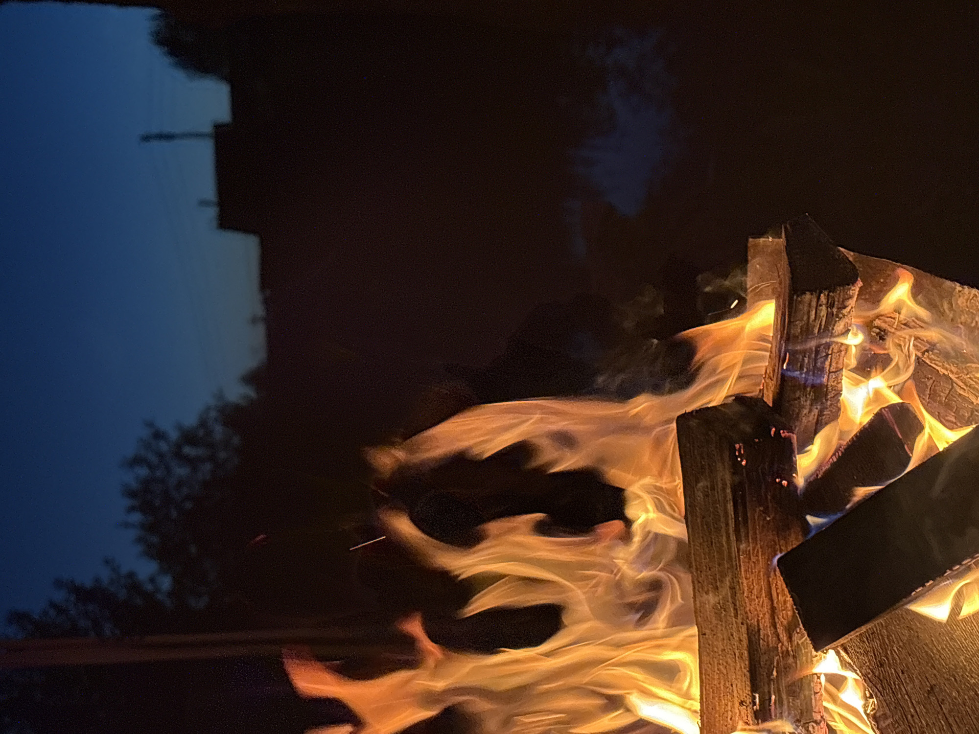 The image shows a campfire burning at dusk or night in an outdoor setting, possibly in the city of Markovo, Russia. The foreground is dominated by logs of wood engulfed in vibrant orange and yellow flames, casting a warm glow. Some embers are visible within the fire. In the background, the sky is a deep twilight blue, transitioning to darker shades, with silhouettes of trees and structures, likely buildings, visible against the horizon. A faint wisps of smoke rise from the fire and dissipate into the dark background. The scene evokes a sense of warmth and a quiet evening outdoors. There are no people or animals visible in the image.